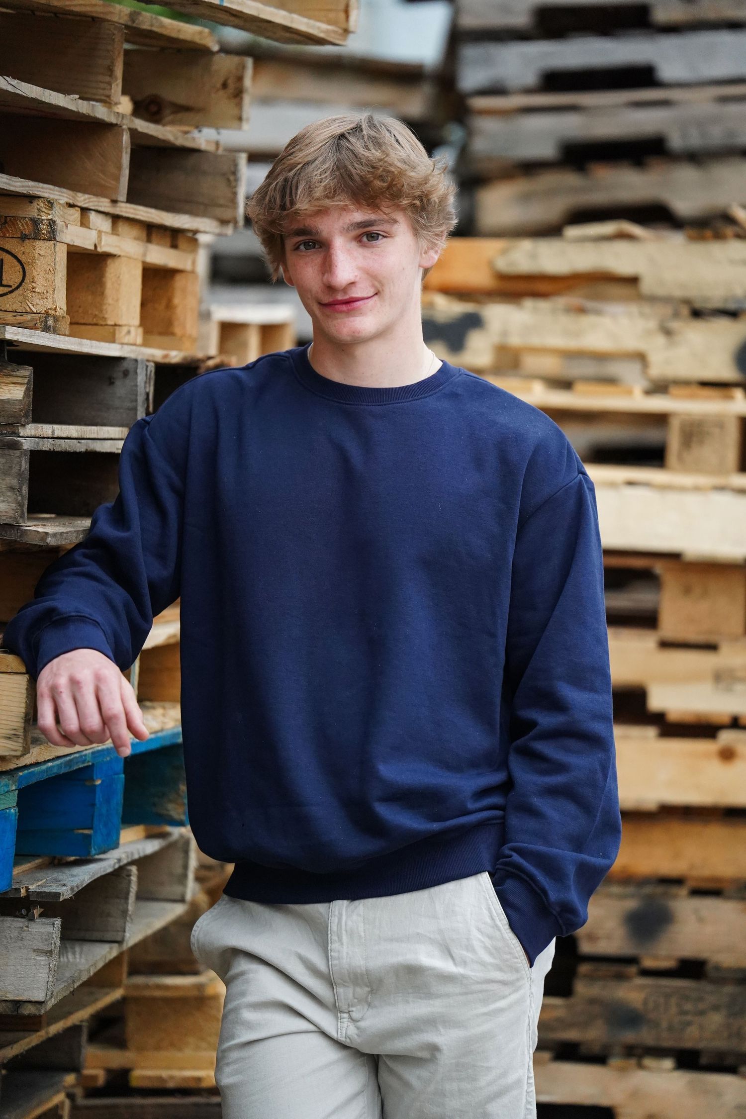 A young person in a navy blue sweatshirt and khaki pants stands in front of stacked wooden pallets.