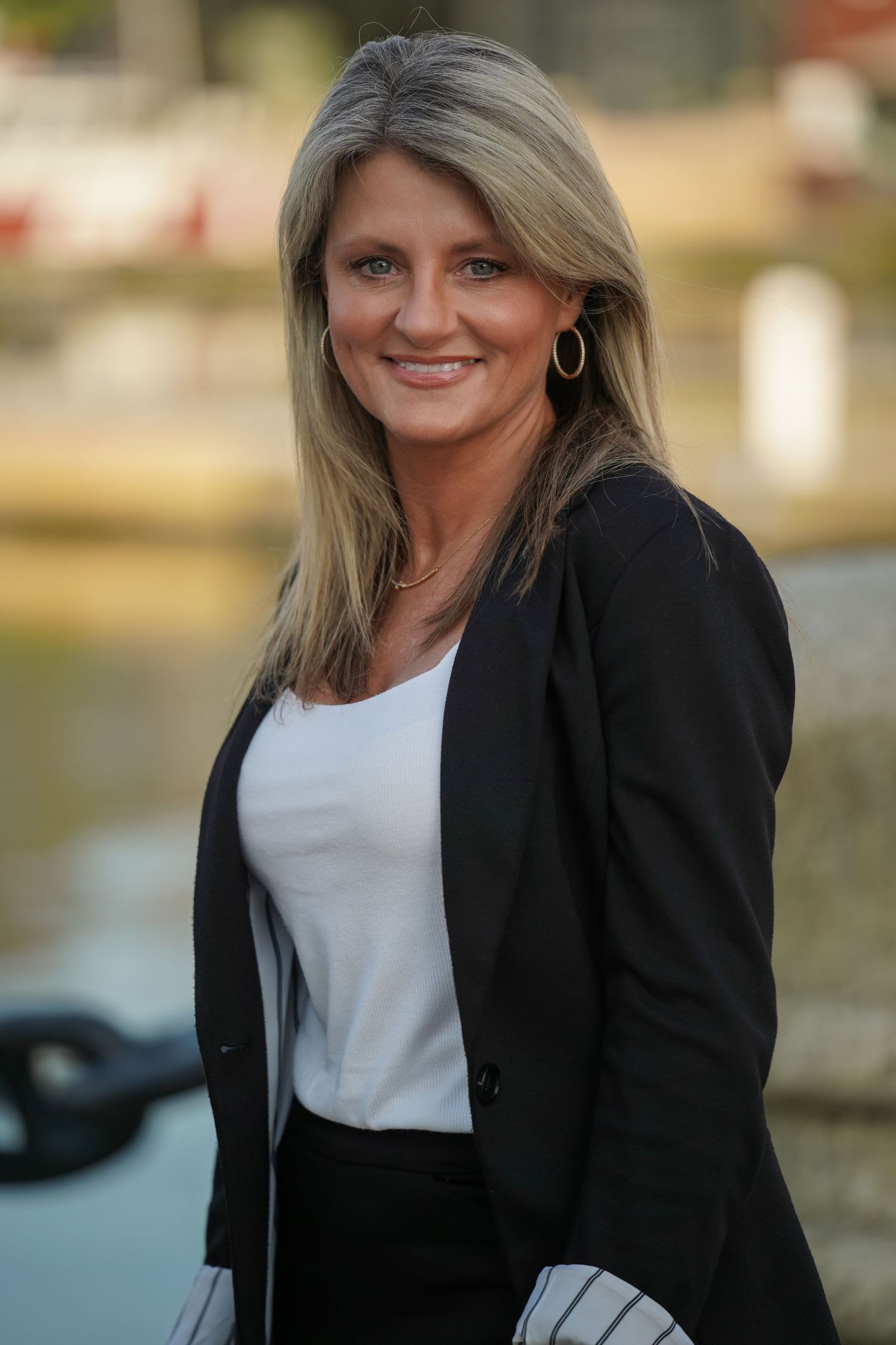 Professional headshot of a person wearing a black blazer and white top against a blurred outdoor background.
