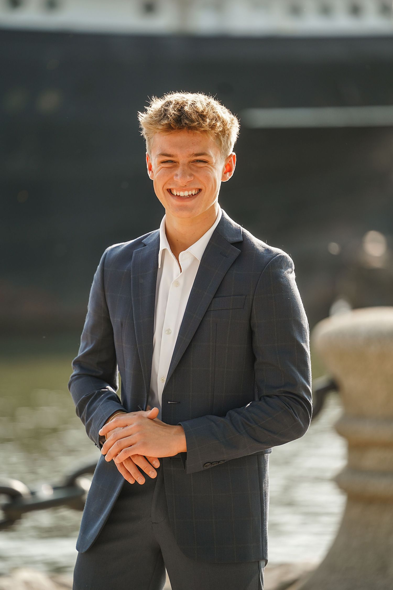 A smiling young person in a dark suit and white dress shirt poses outdoors in natural sunlight.
