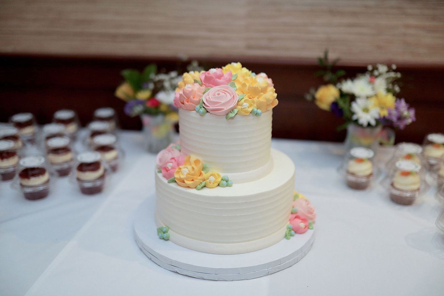 An elegant white wedding cake decorated with pastel flowers sits next to rows of dessert cups on a wooden table.
