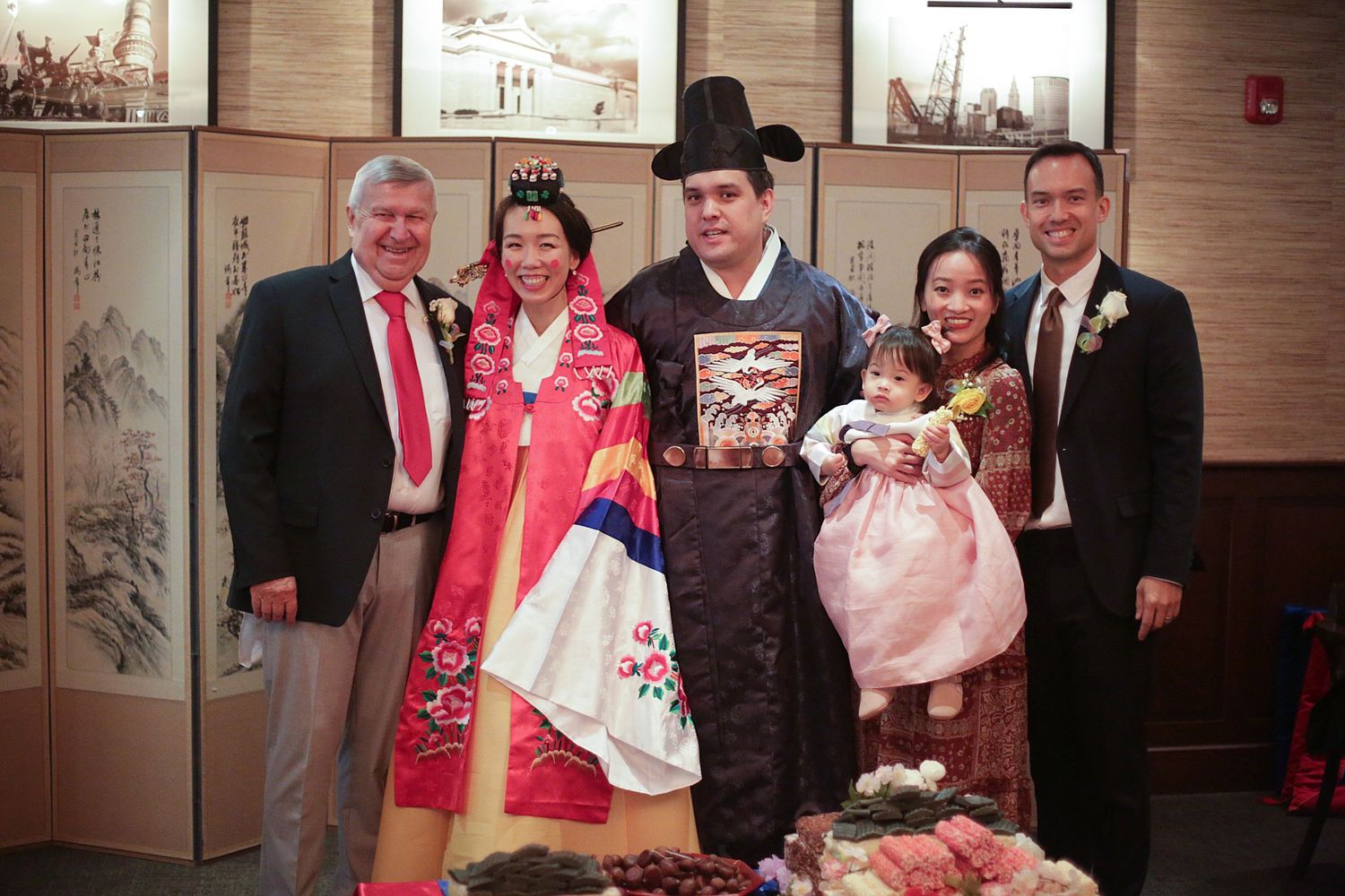 A group in traditional Korean hanbok and formal attire pose together during a celebration at an Asian restaurant interior.