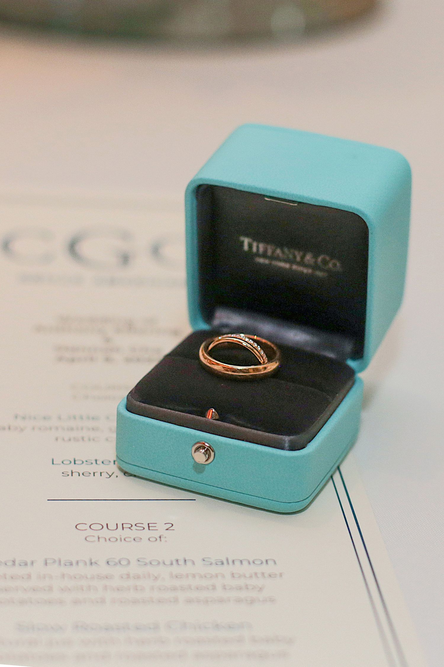 A gold ring sits in an iconic Tiffany & Co. turquoise blue box atop a printed menu or document.