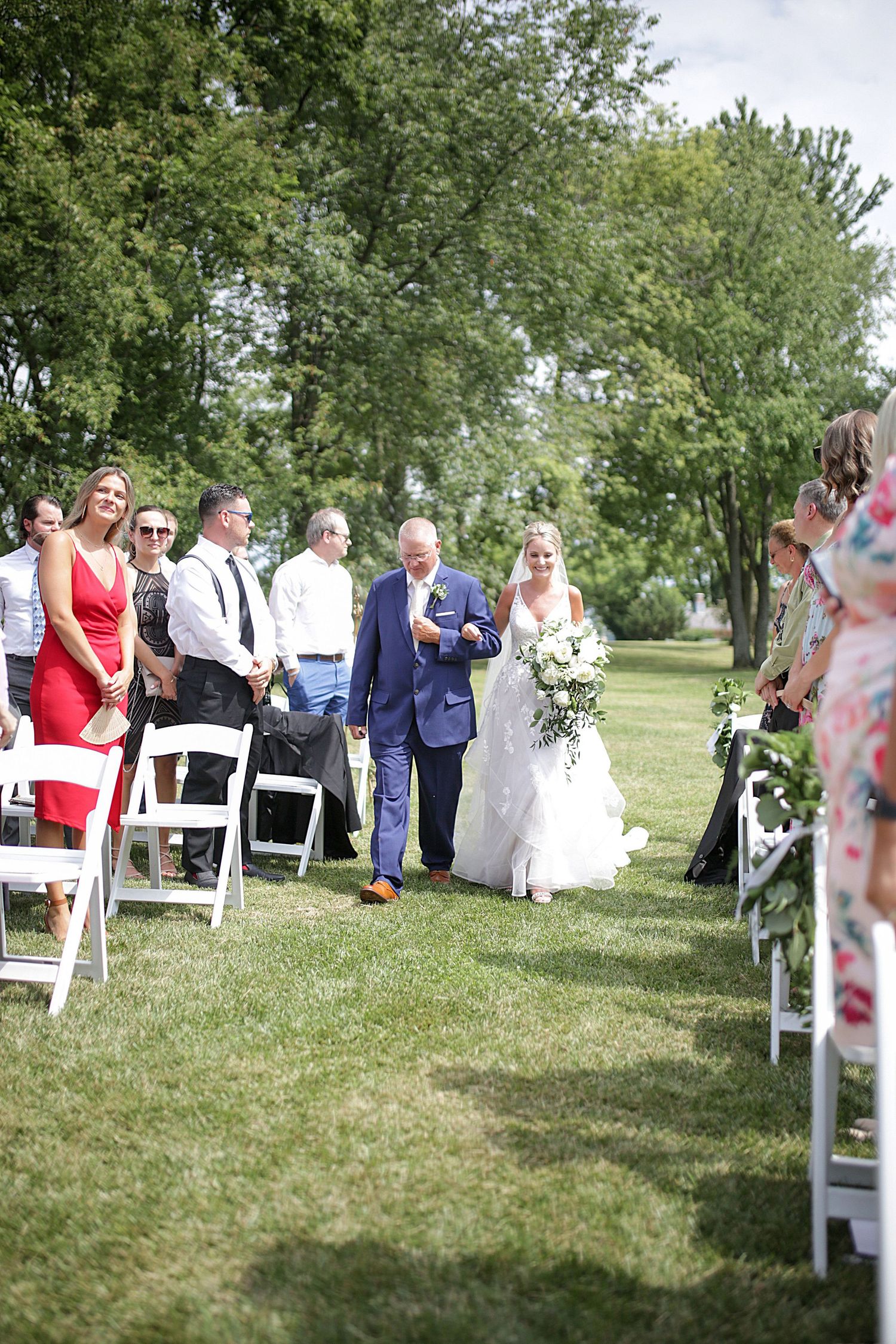 Outdoor wedding ceremony processional on a grassy lawn lined with white chairs and floral decorations.