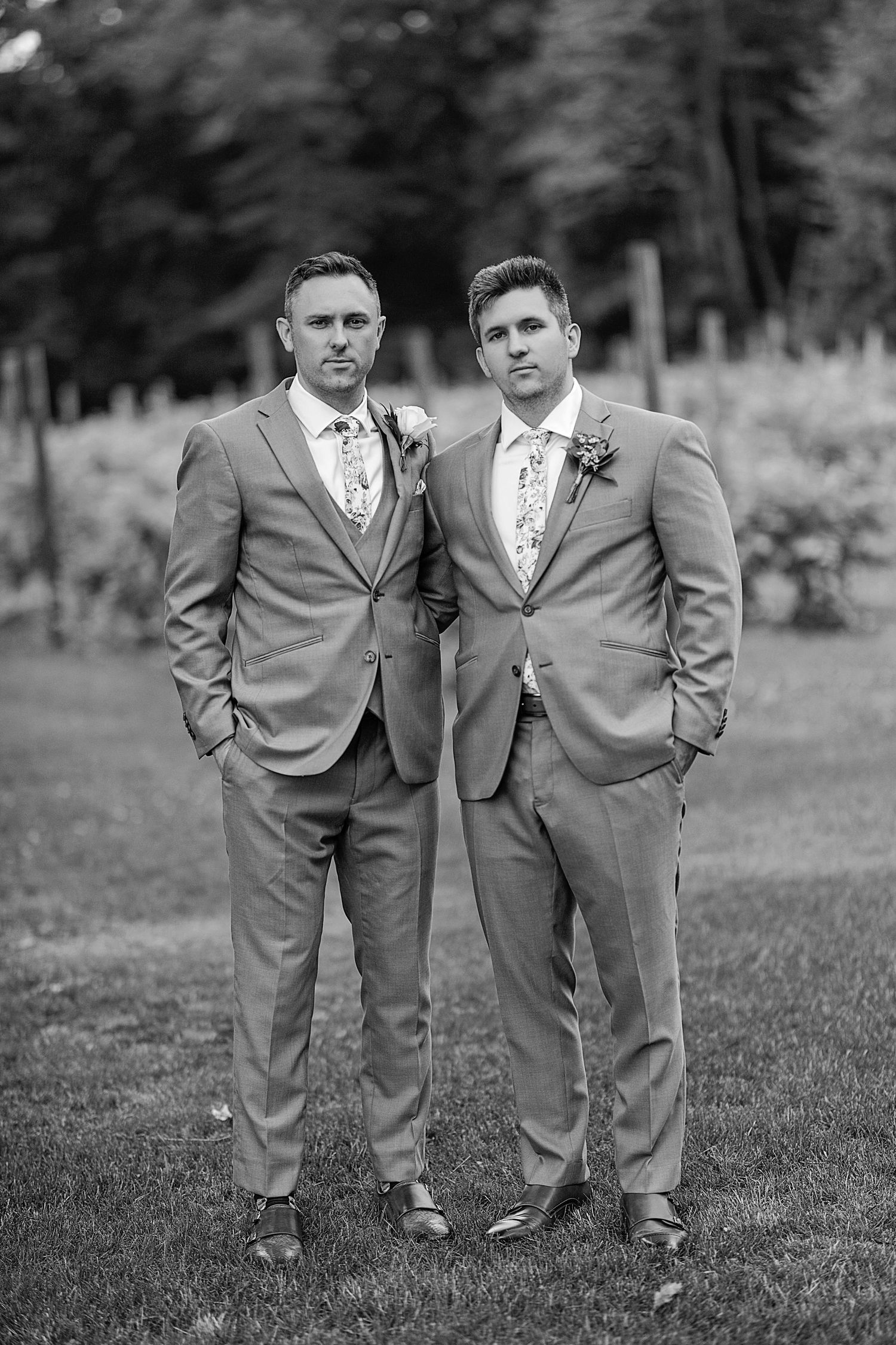 Two suited figures pose formally in grey wedding attire against a natural backdrop.