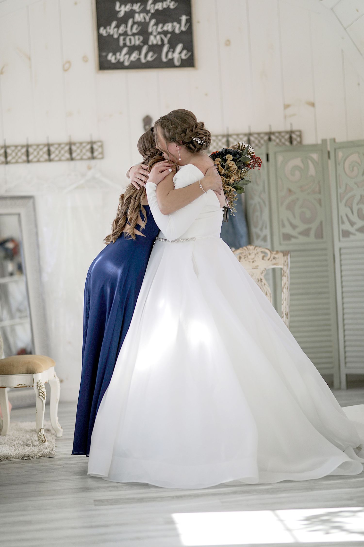 Two wedding attendees share an emotional embrace, with one in a flowing white gown and the other in navy blue.