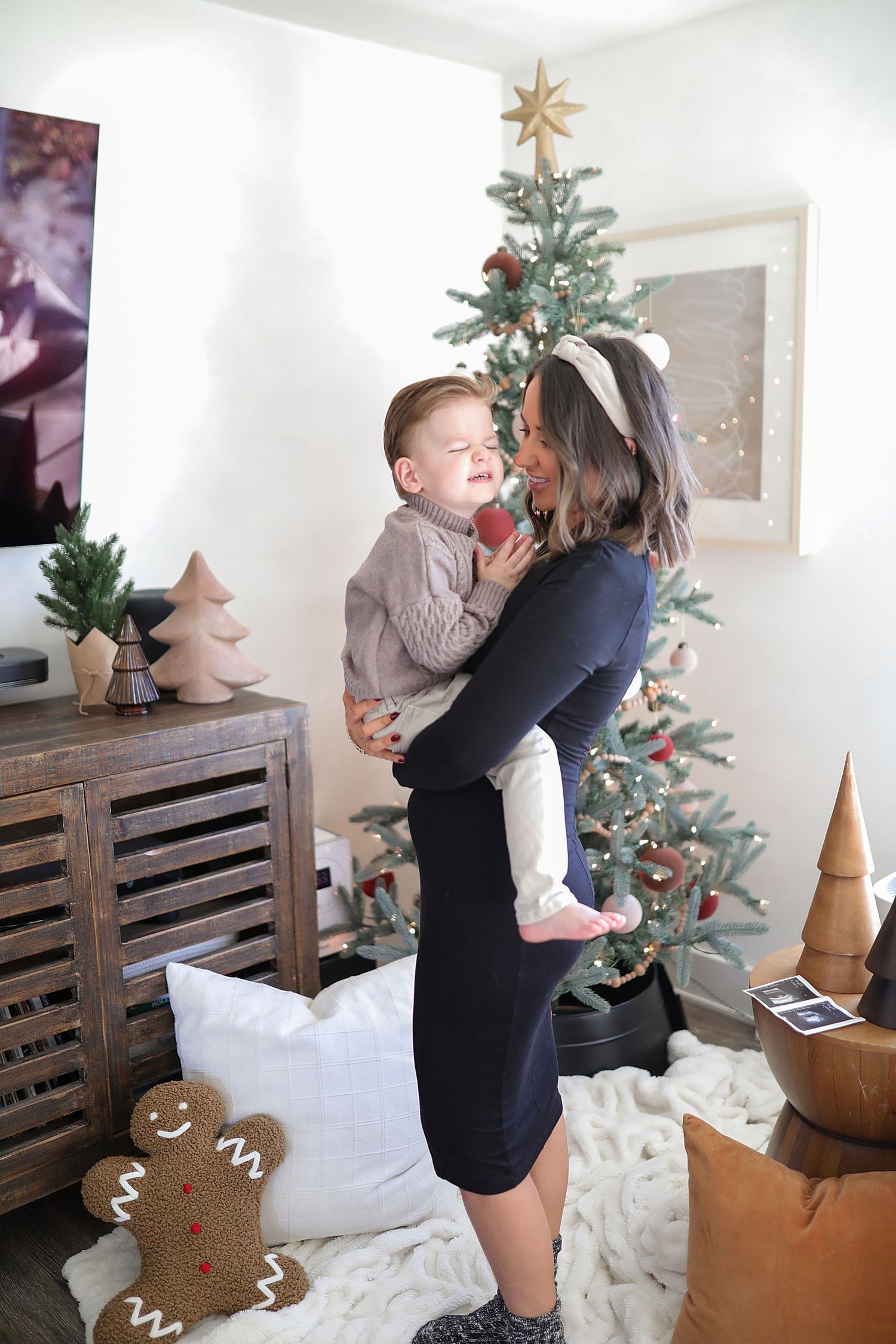 Woman in black dress dancing with toddler in front of decorated Christmas tree in cozy home setting.