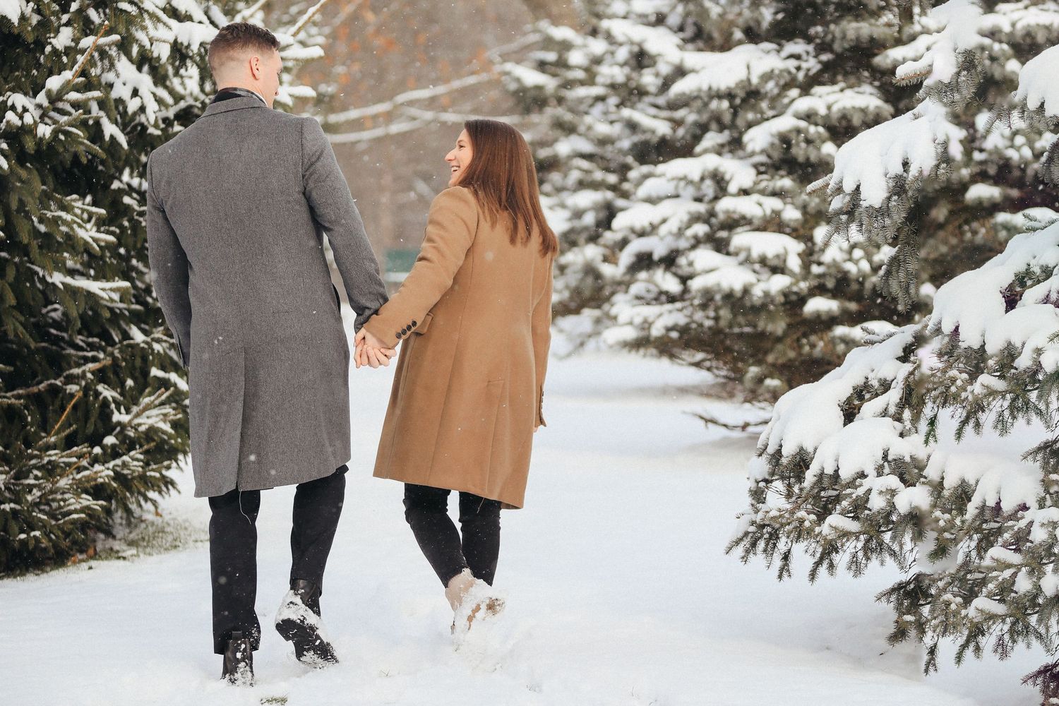 A couple walks hand in hand through a snowy winter path surrounded by evergreen trees in a romantic setting.