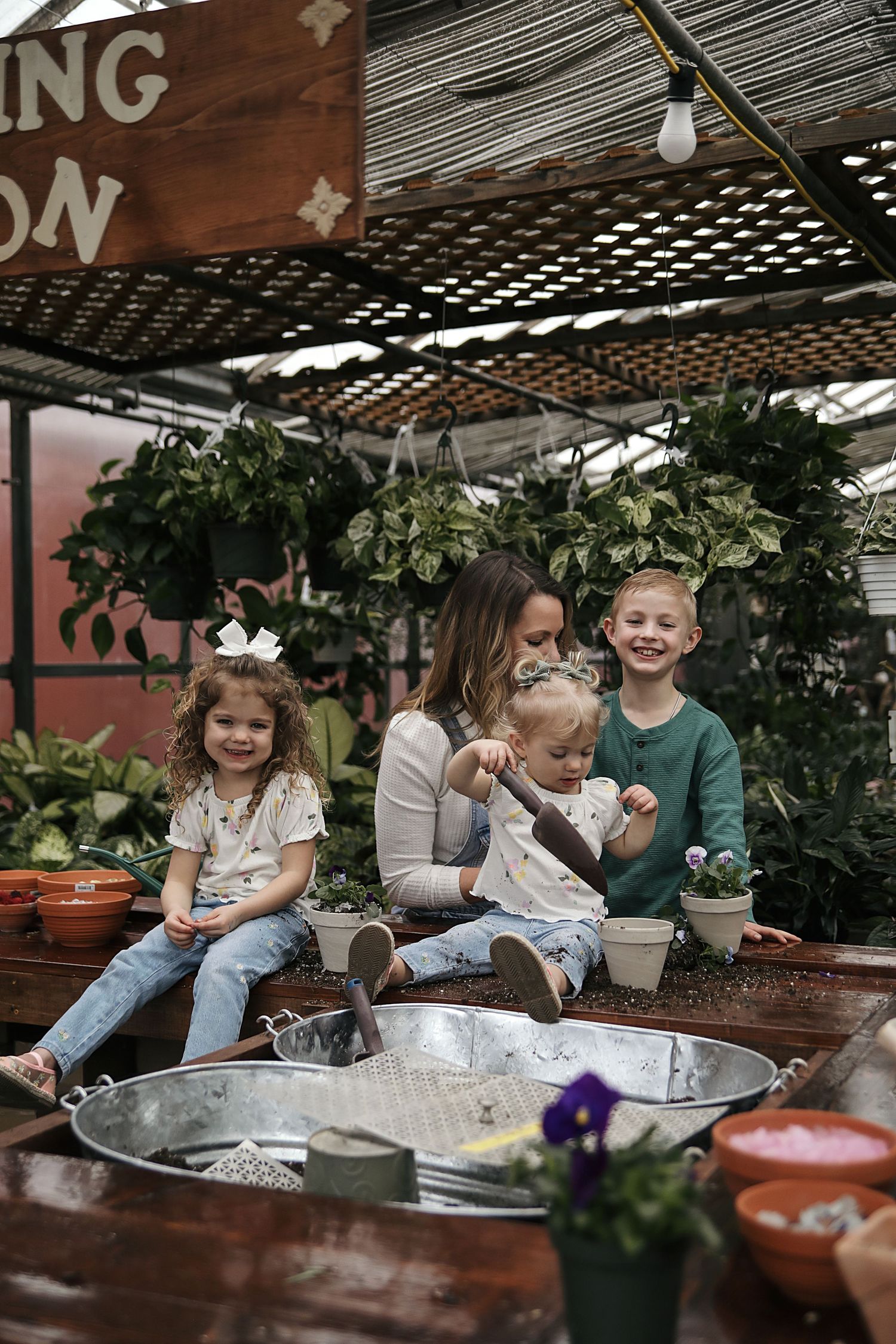 Tea party in a greenhouse setting with children and adults gathered around a table with plants in the background.