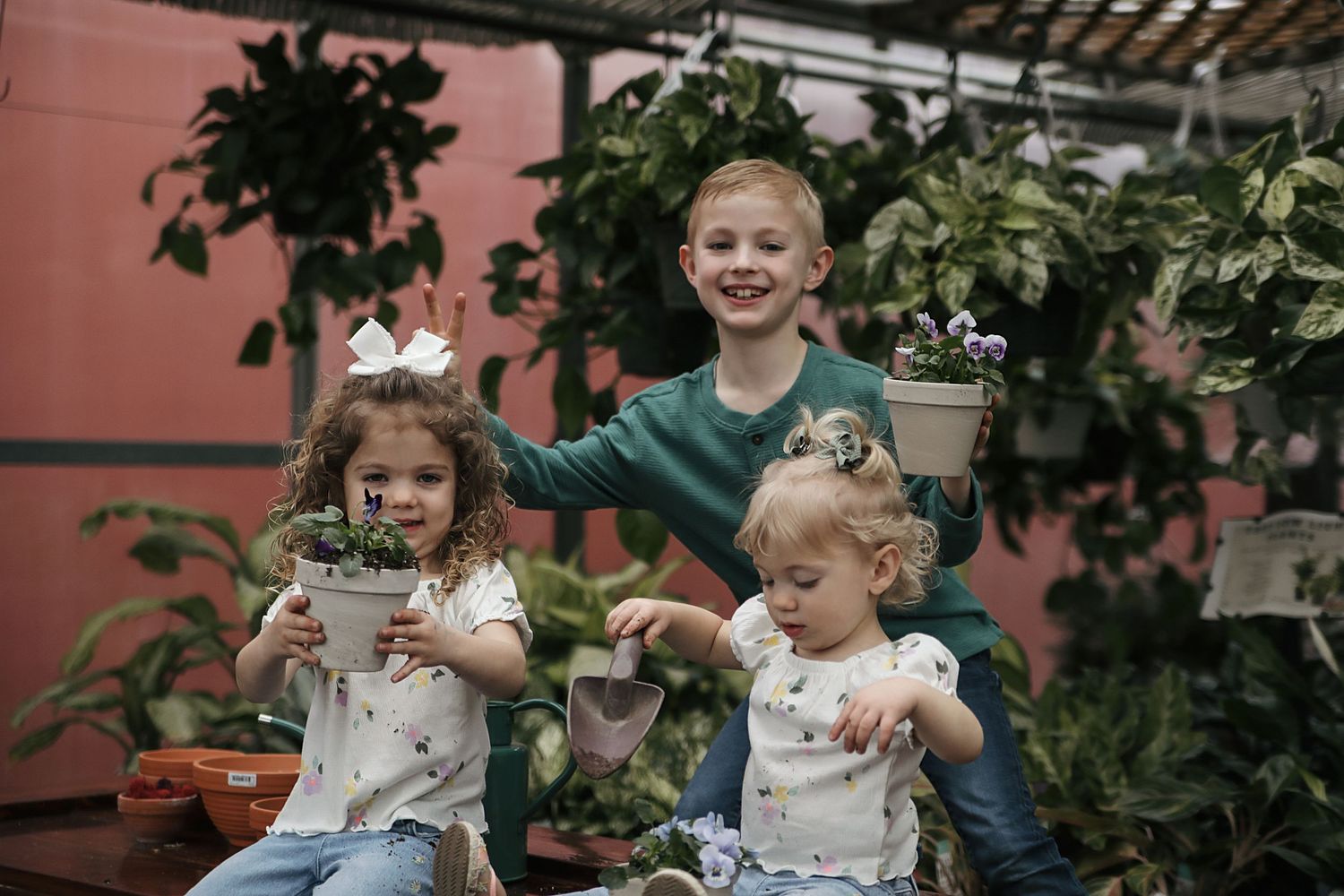 Three children sit together surrounded by potted plants and greenery in a rustic greenhouse setting.