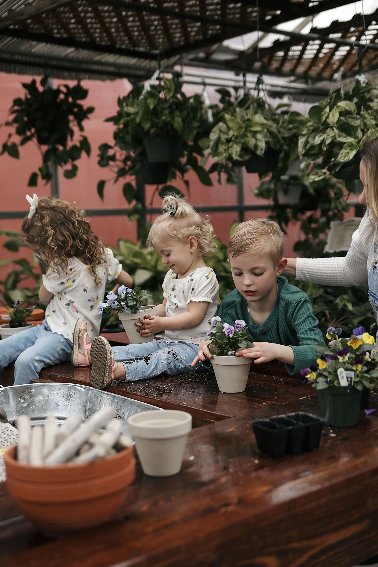 Children gather around a wooden table in a greenhouse setting while planting and potting flowers together.