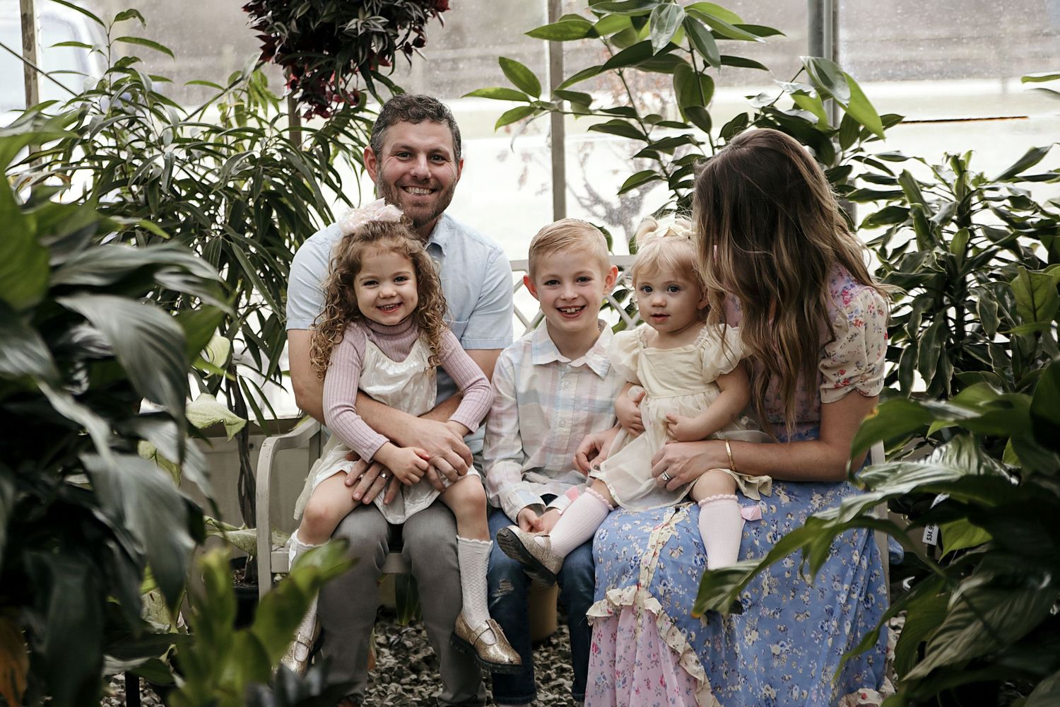 A cheerful group sits together surrounded by lush green plants in a greenhouse setting.
