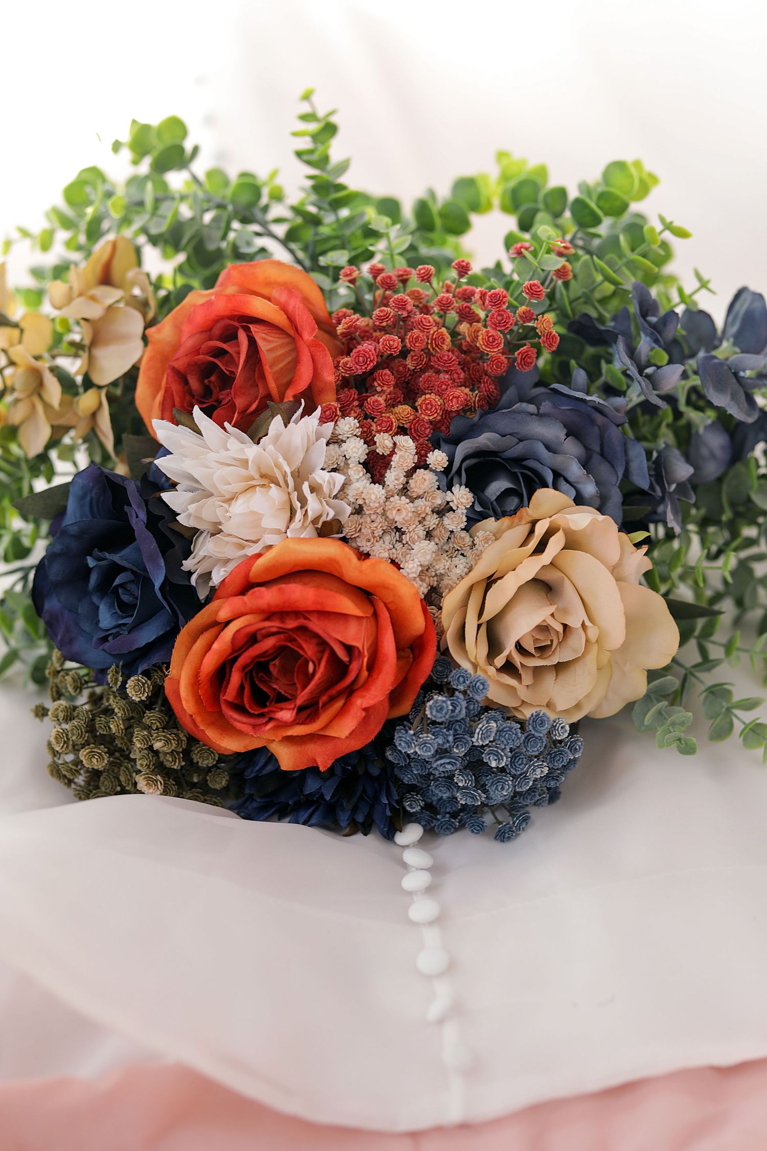 Wedding bouquet with orange roses, navy blue flowers, and greenery on white background.