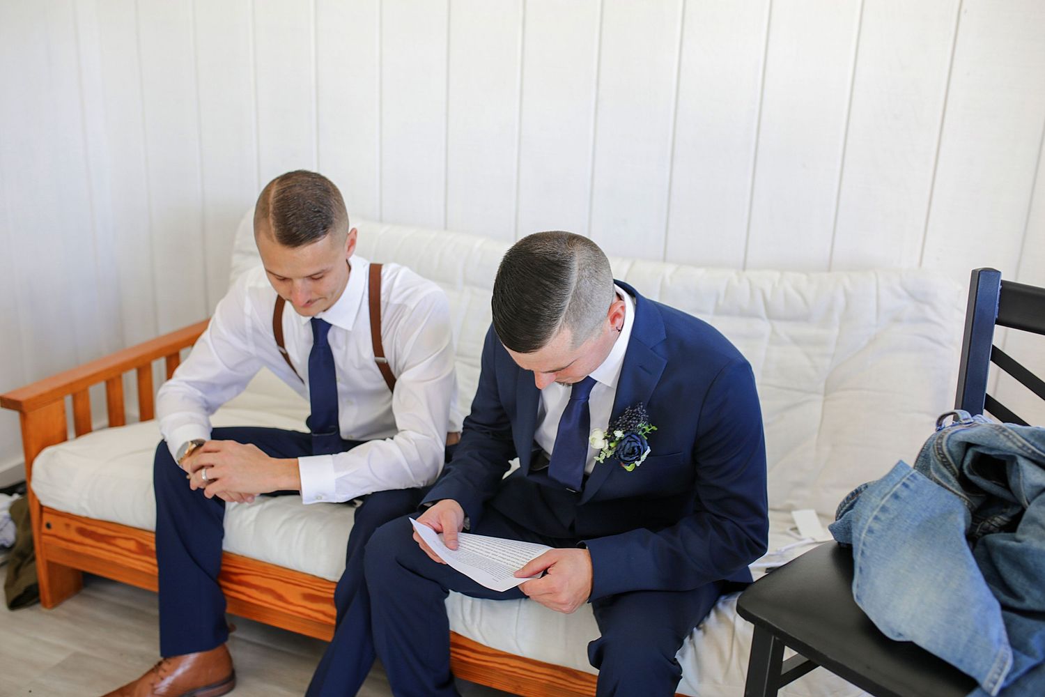 Two men in suits and ties sit on a wooden bench while reading papers before an event.