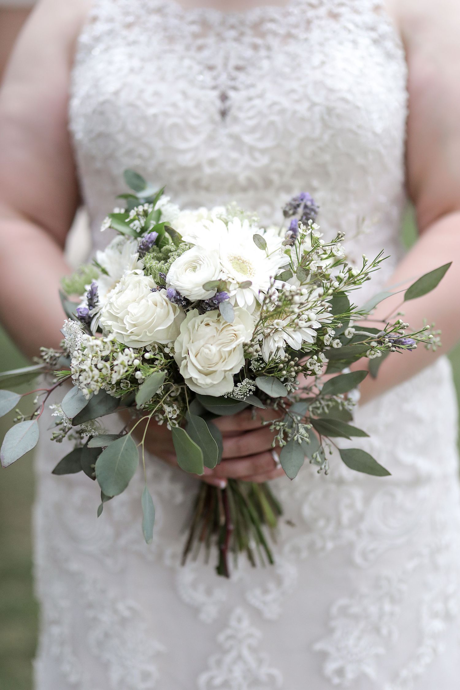 Close-up of white wedding bouquet with roses, eucalyptus leaves, and delicate purple accents held against lace dress.