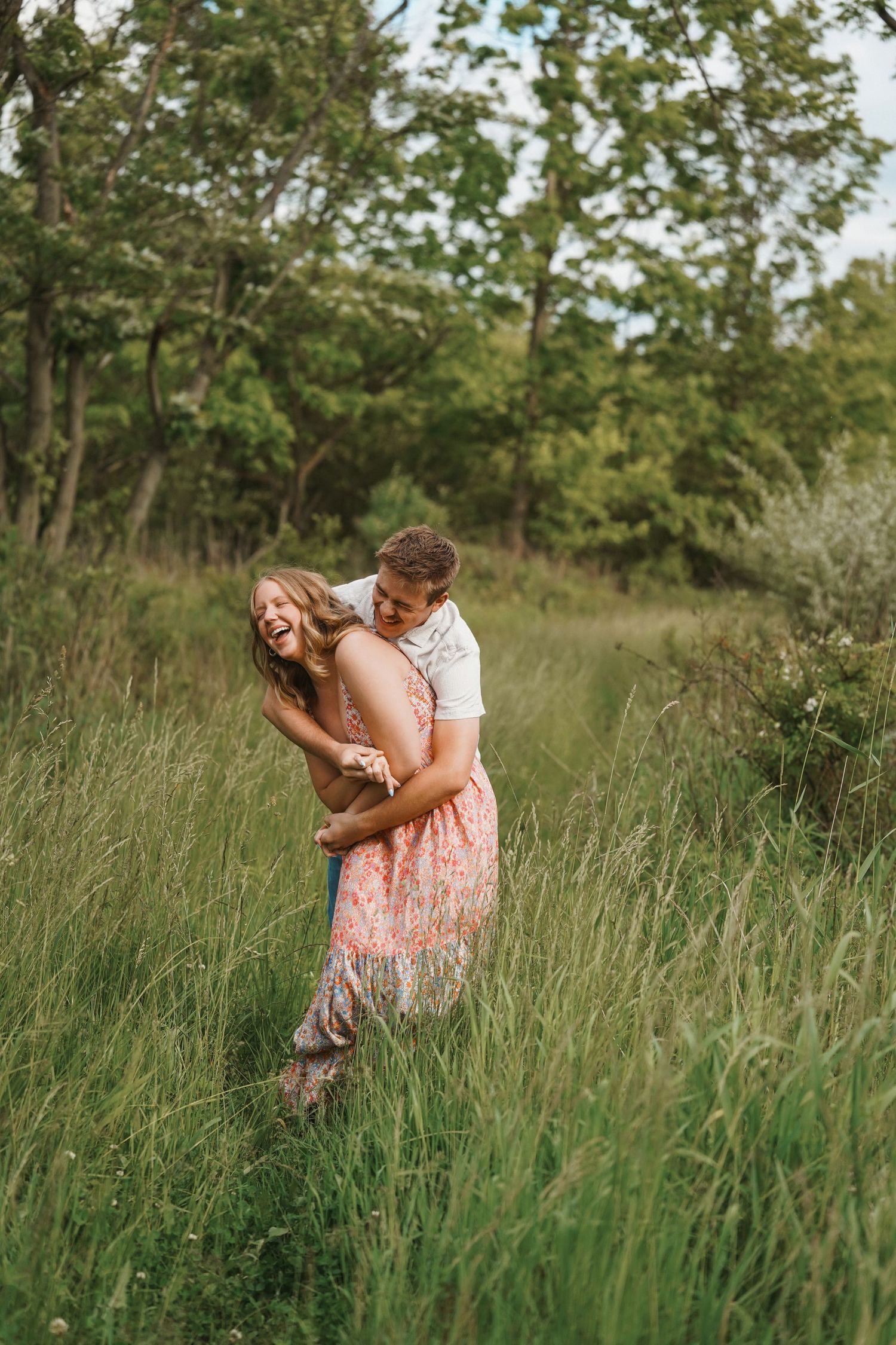 A loving couple embraces and laughs together in a field of tall grass with green trees in the background.