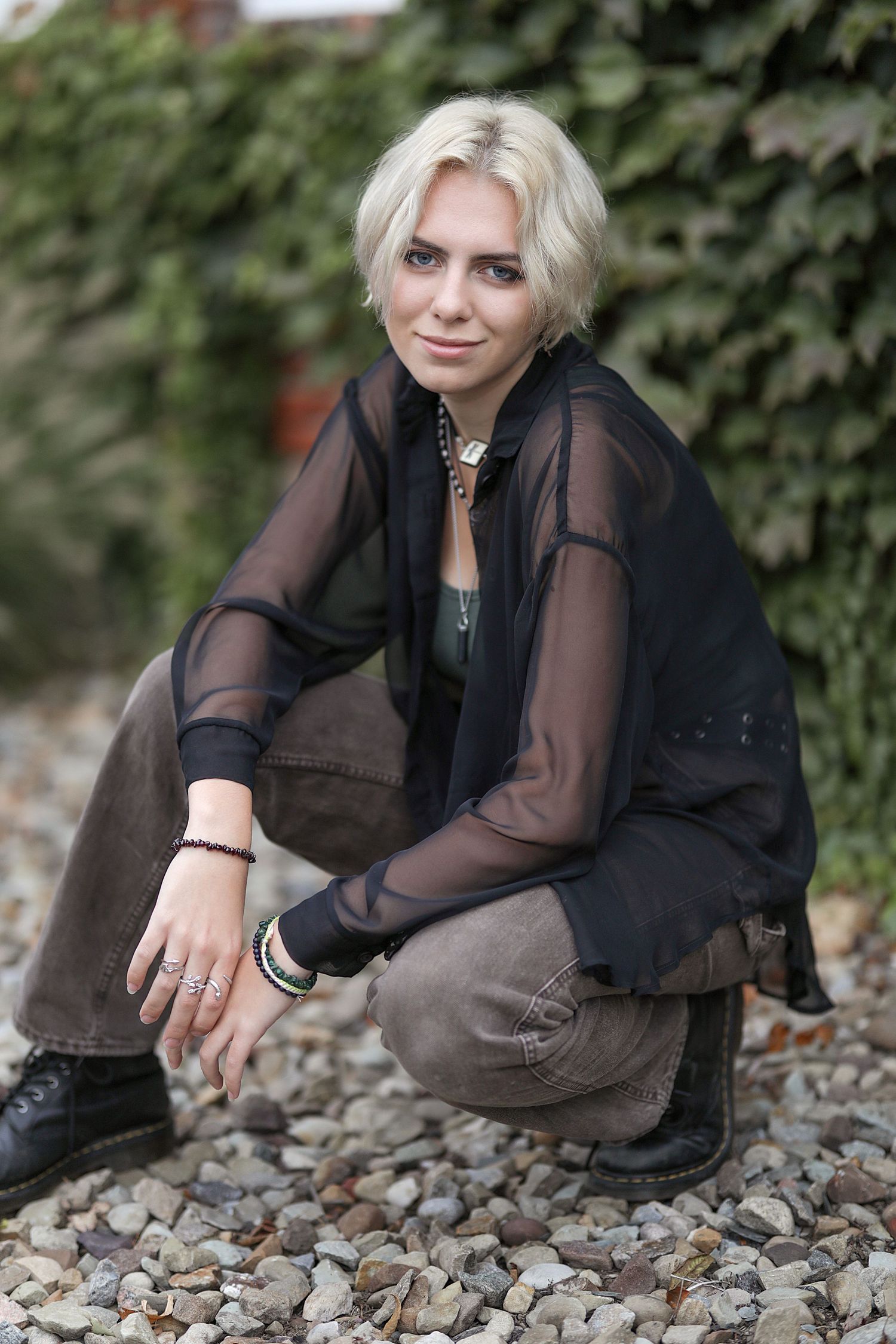 Person in sheer black top and brown pants sits casually on stone ground with green ivy backdrop.