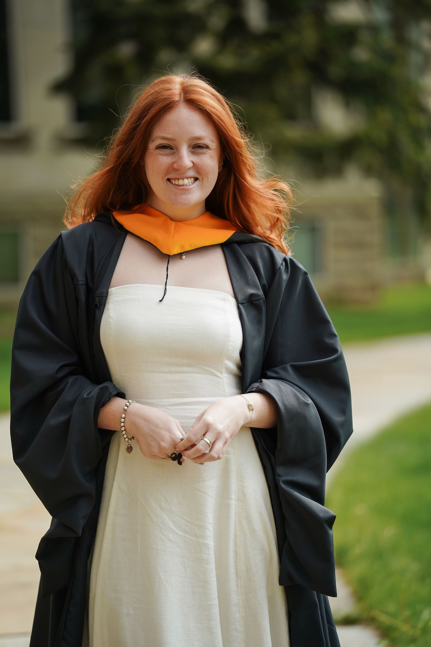 Graduate wearing black academic regalia and white dress poses outdoors with bright orange hood for commencement photo.