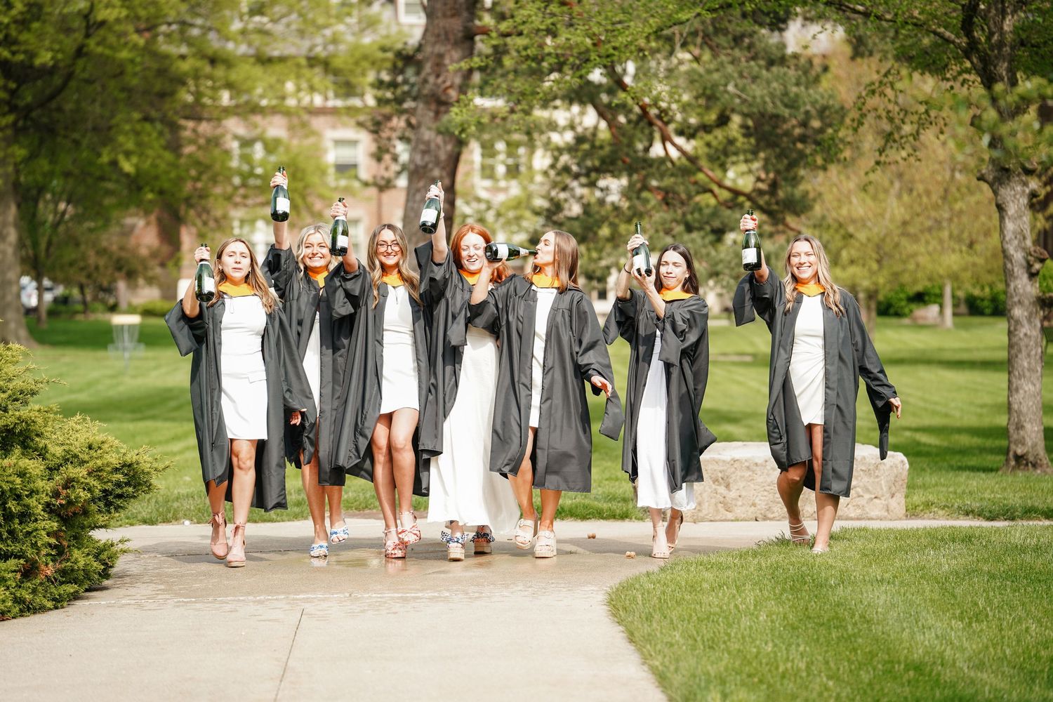 A group in graduation caps and gowns walks together on a sunny green campus path celebrating commencement.