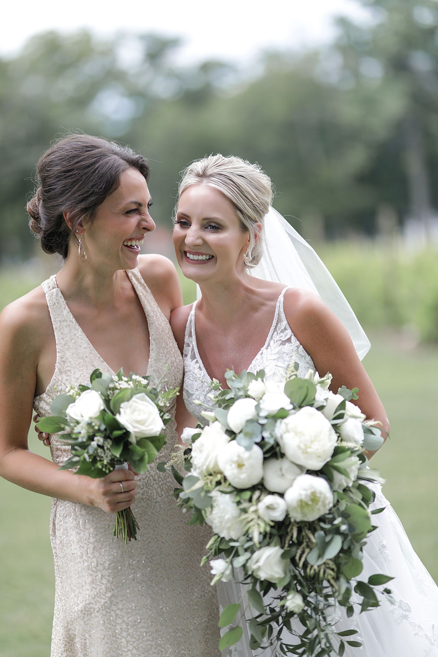 Two wedding party members hold white floral bouquets while sharing a joyful moment outdoors.