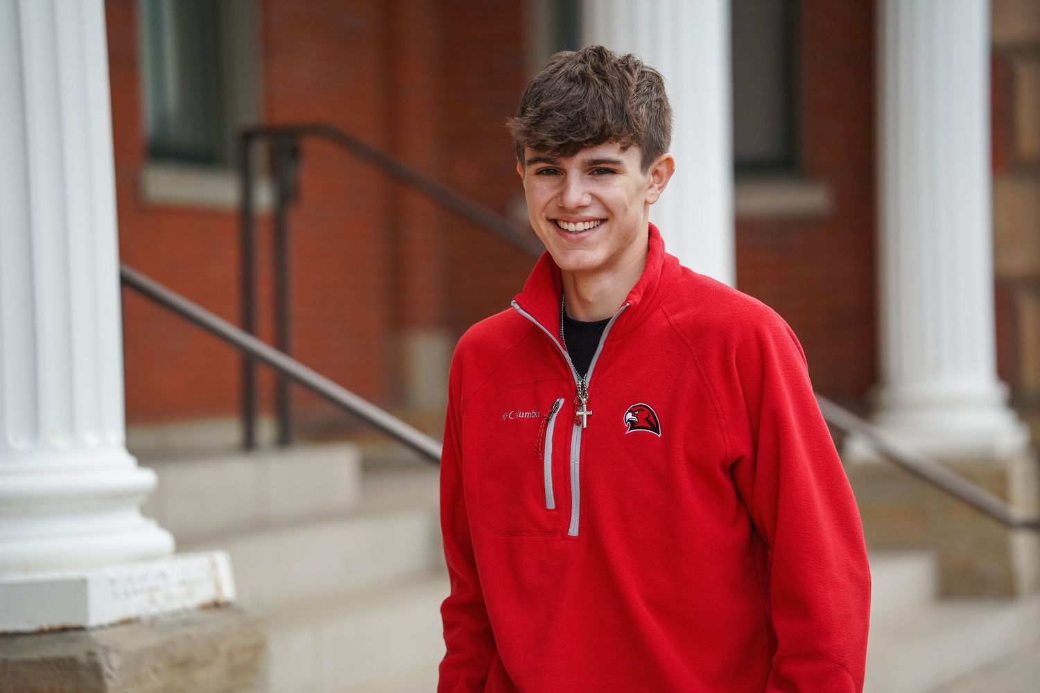 A smiling student in a red quarter-zip pullover stands outside a brick building with white columns.