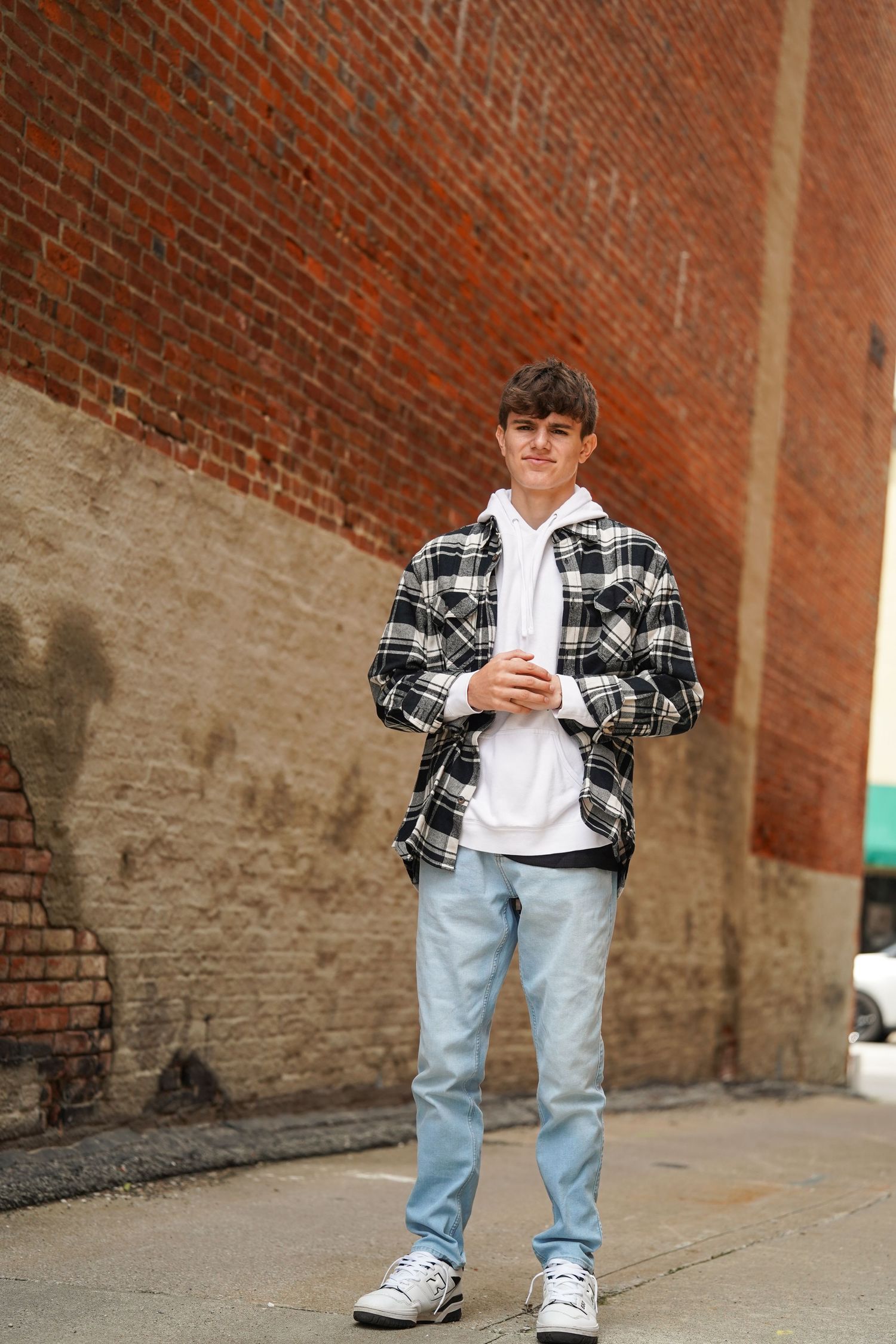 Person standing against brick wall wearing plaid flannel jacket, white t-shirt, light jeans and sneakers.