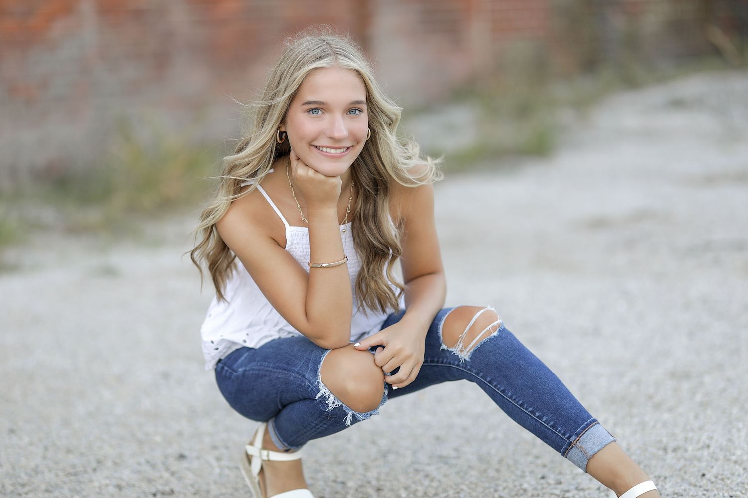 A smiling figure wearing blue jeans and white shoes sits casually on the ground outdoors.