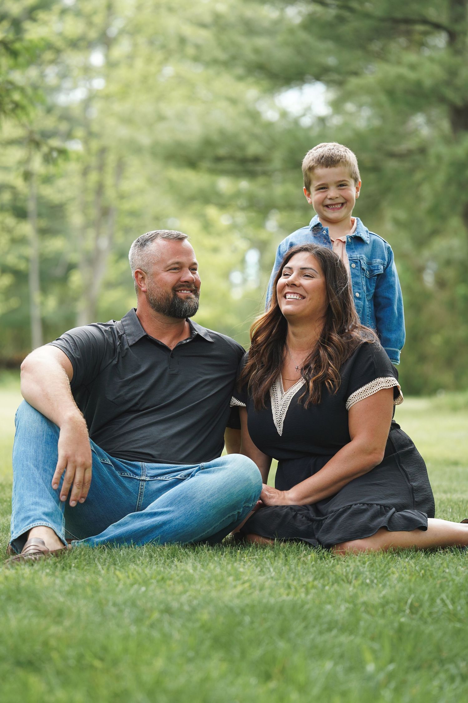 Three family members sit together on green grass in a sun-dappled outdoor setting for a casual portrait.