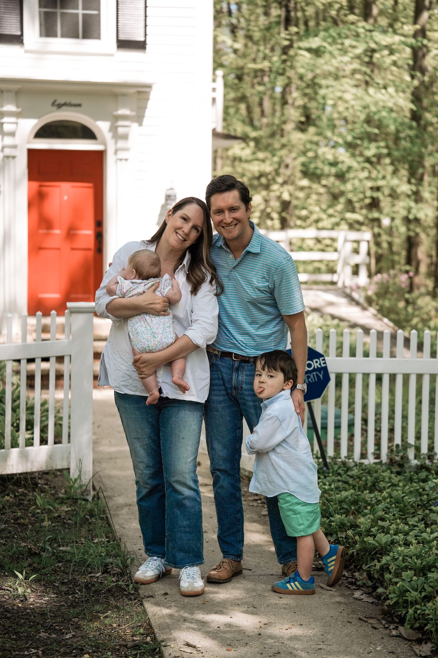 Family standing outside of house with red door and white picket fence in garden setting.