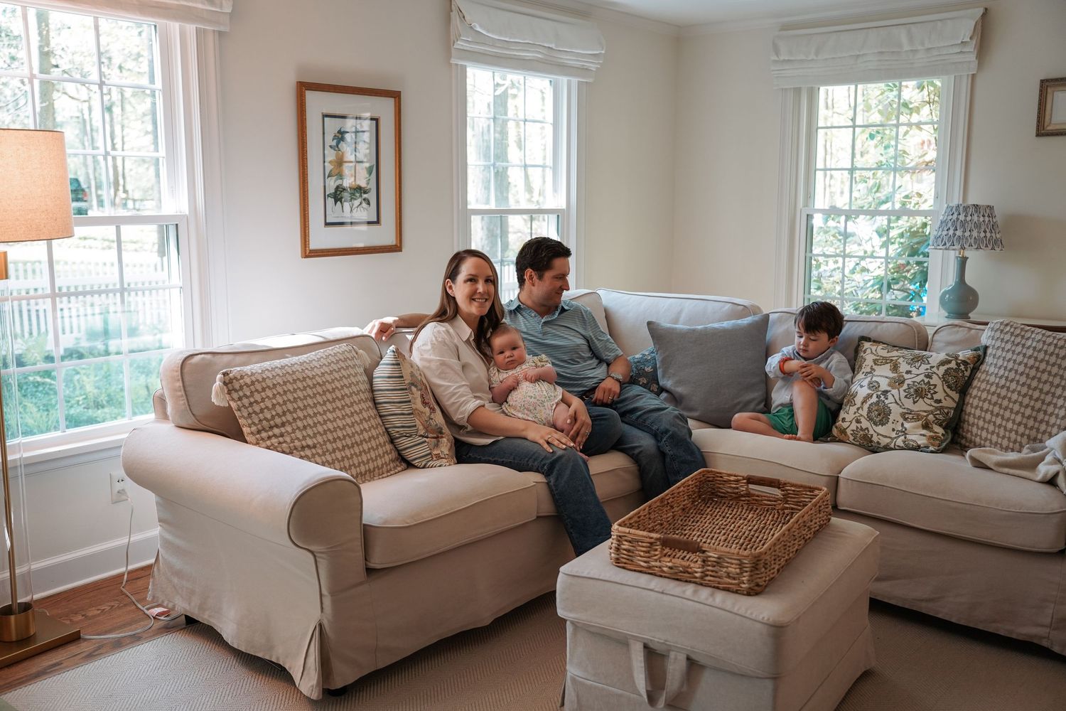 Family relaxes together on beige sectional couch in bright sunlit living room with white decor.