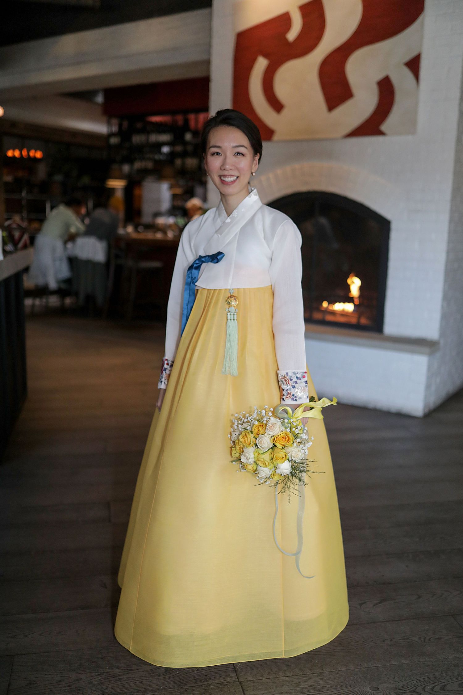 A person wearing a traditional Korean hanbok with a white top and yellow skirt holds a floral bouquet in an indoor setting.