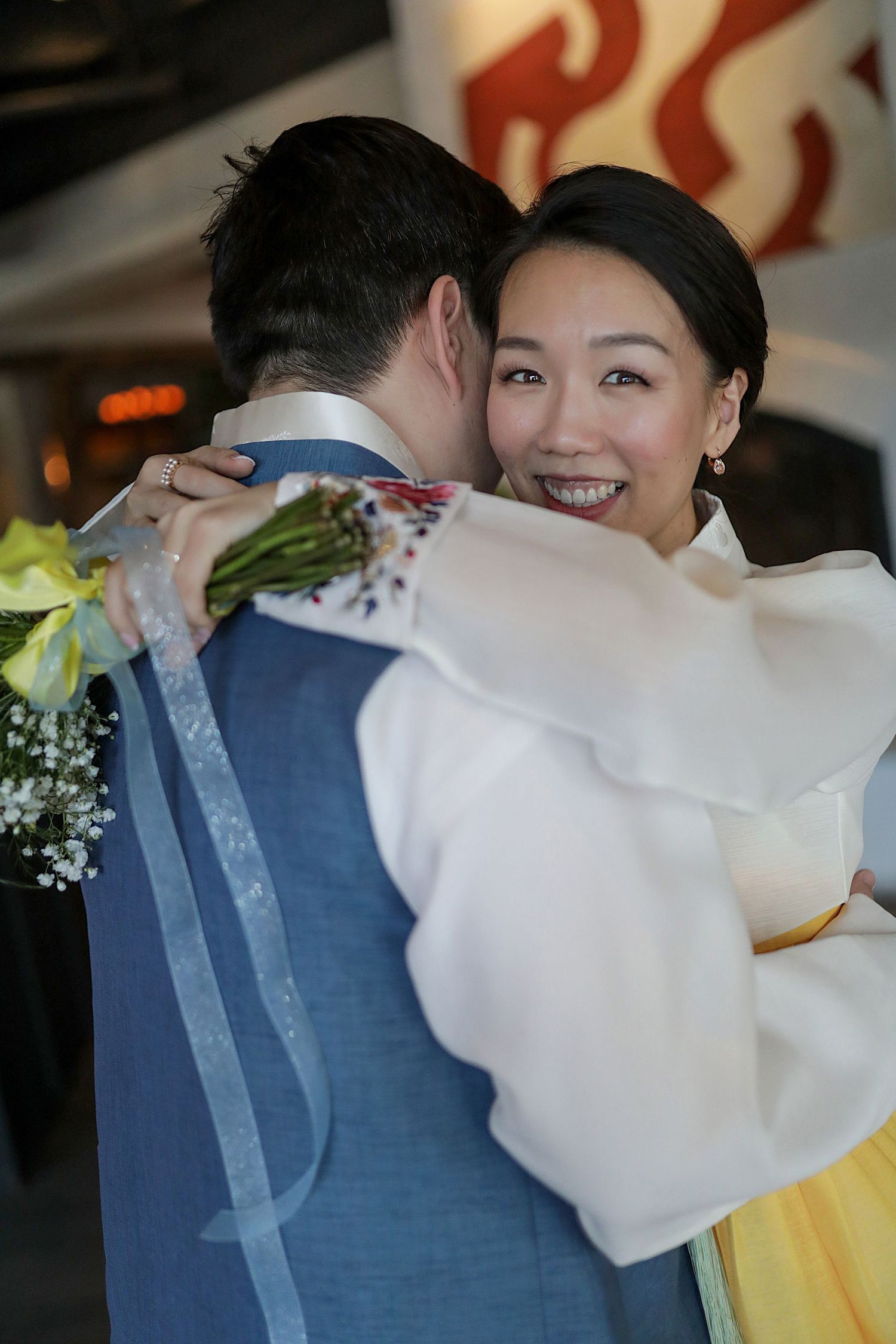 A joyful embrace at a wedding celebration, with a white dress and navy suit visible against decorative background lighting.