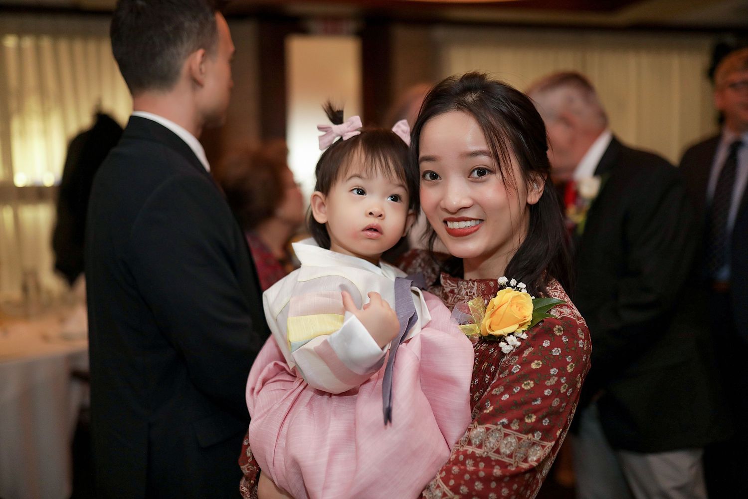 At a formal event, a woman in a burgundy dress holds a baby dressed in pink with a hair bow.