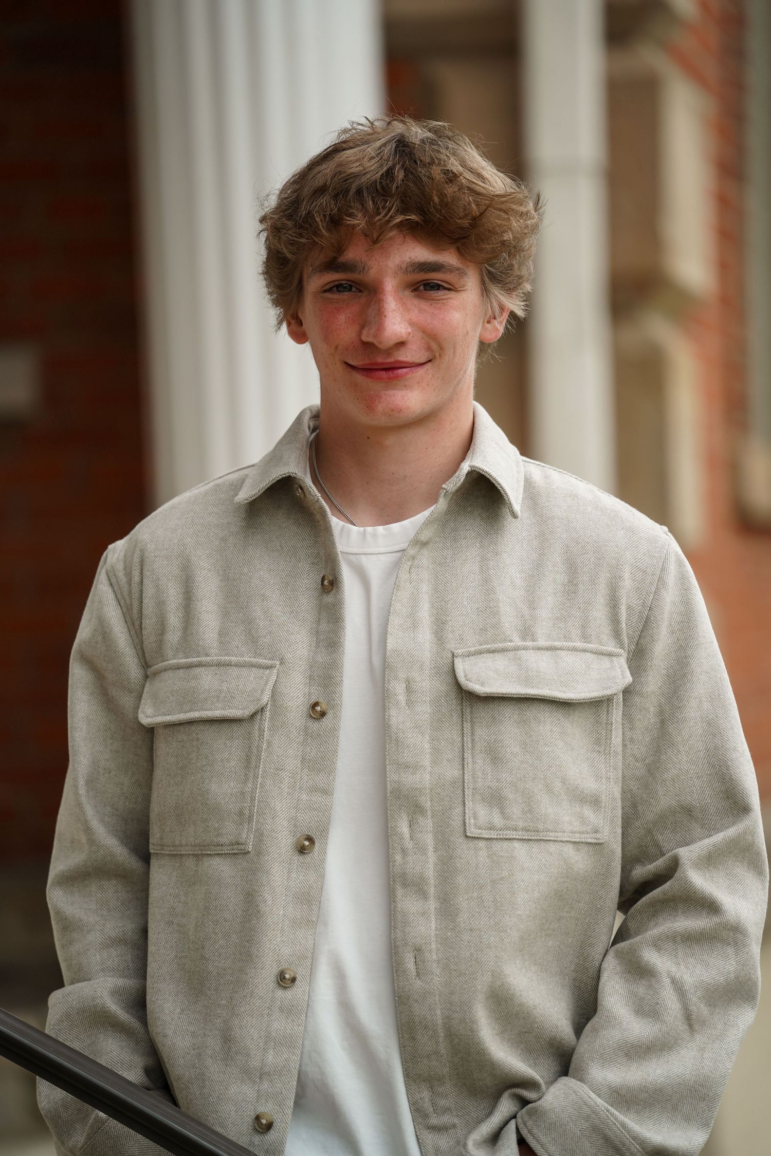 Person wearing a light beige jacket and white t-shirt poses against a columned backdrop.