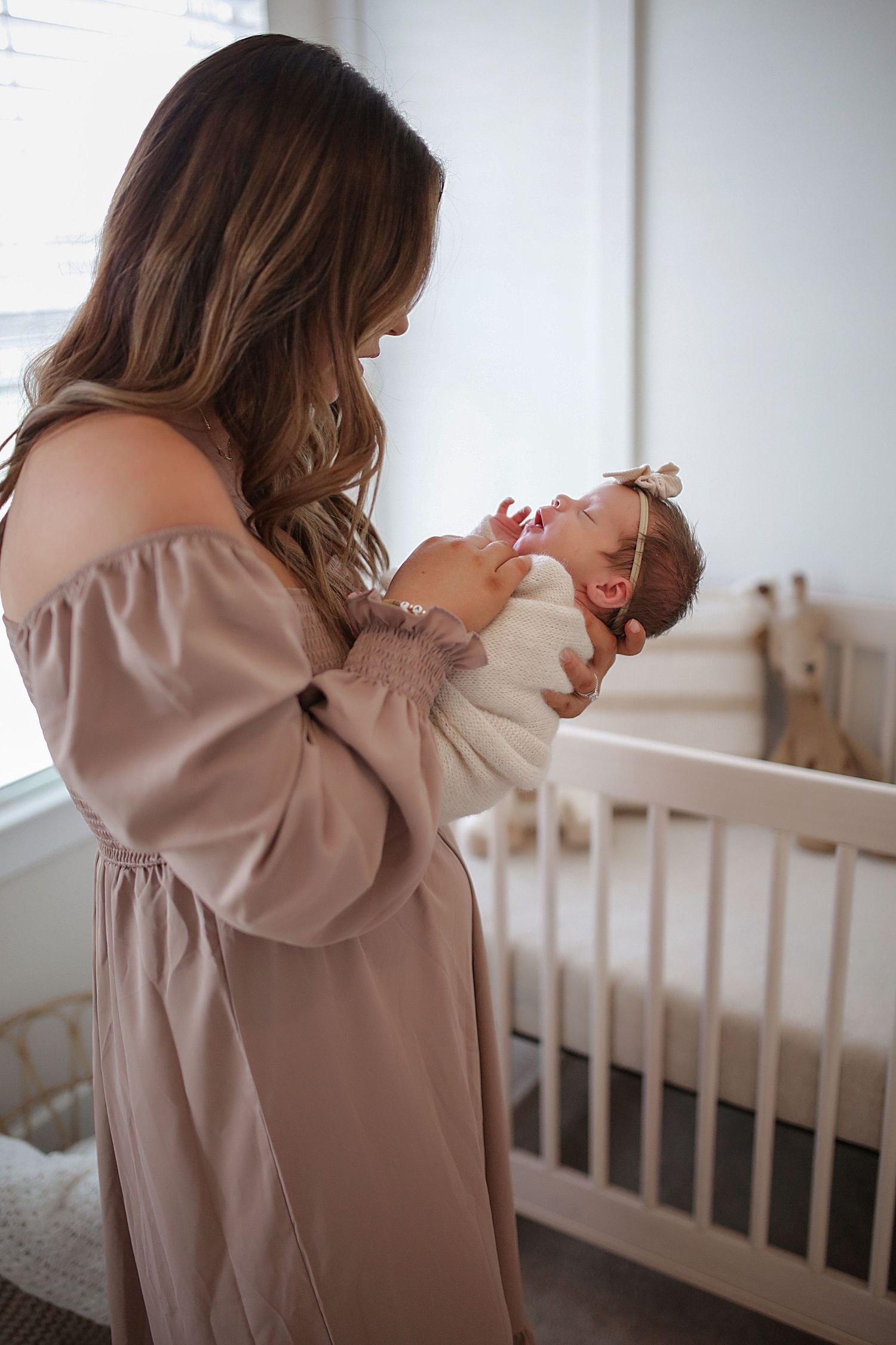A tender moment in a nursery as parent holds newborn baby near white crib, wearing soft mauve dress.