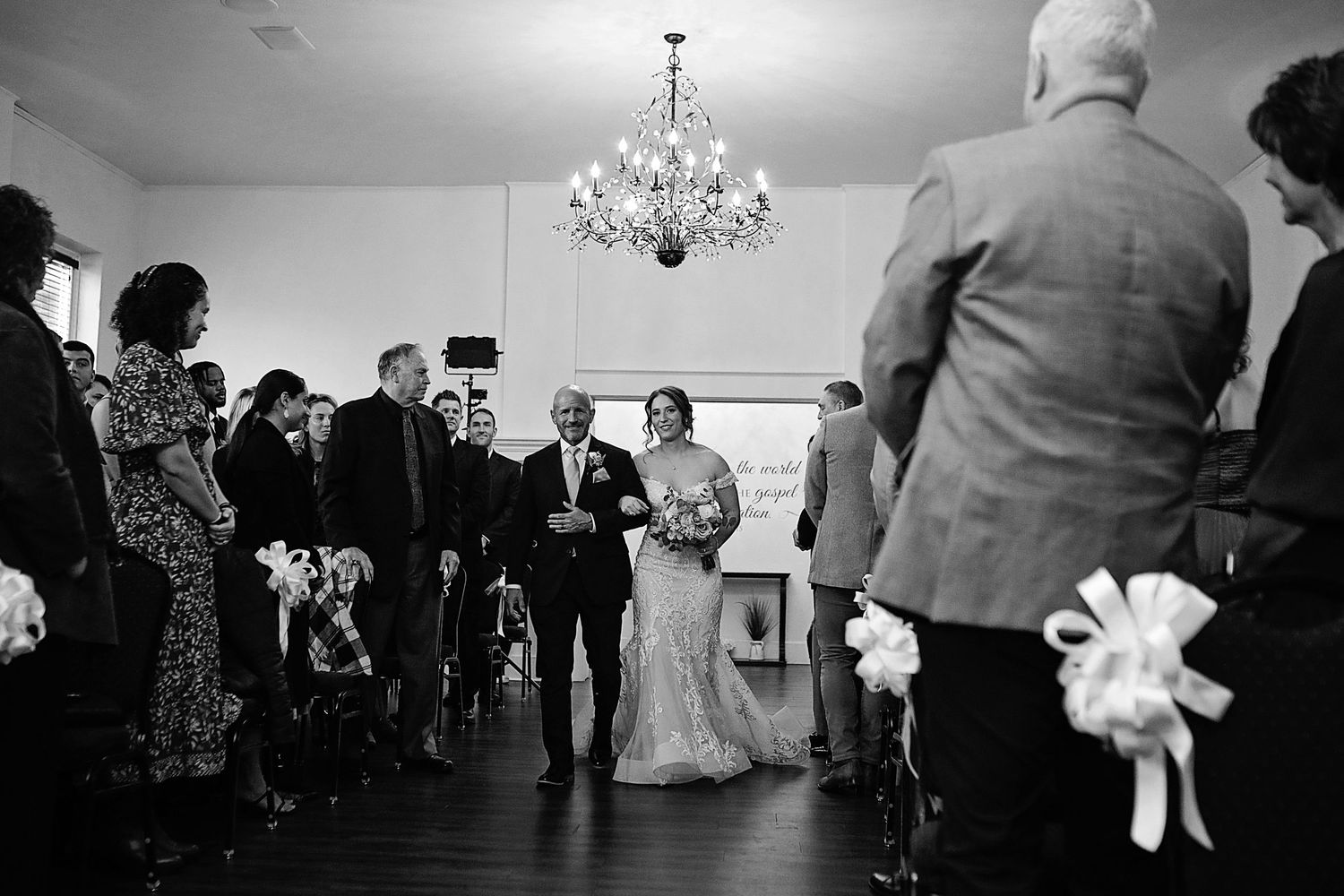 Wedding reception guests dance together under a chandelier during an indoor celebration.