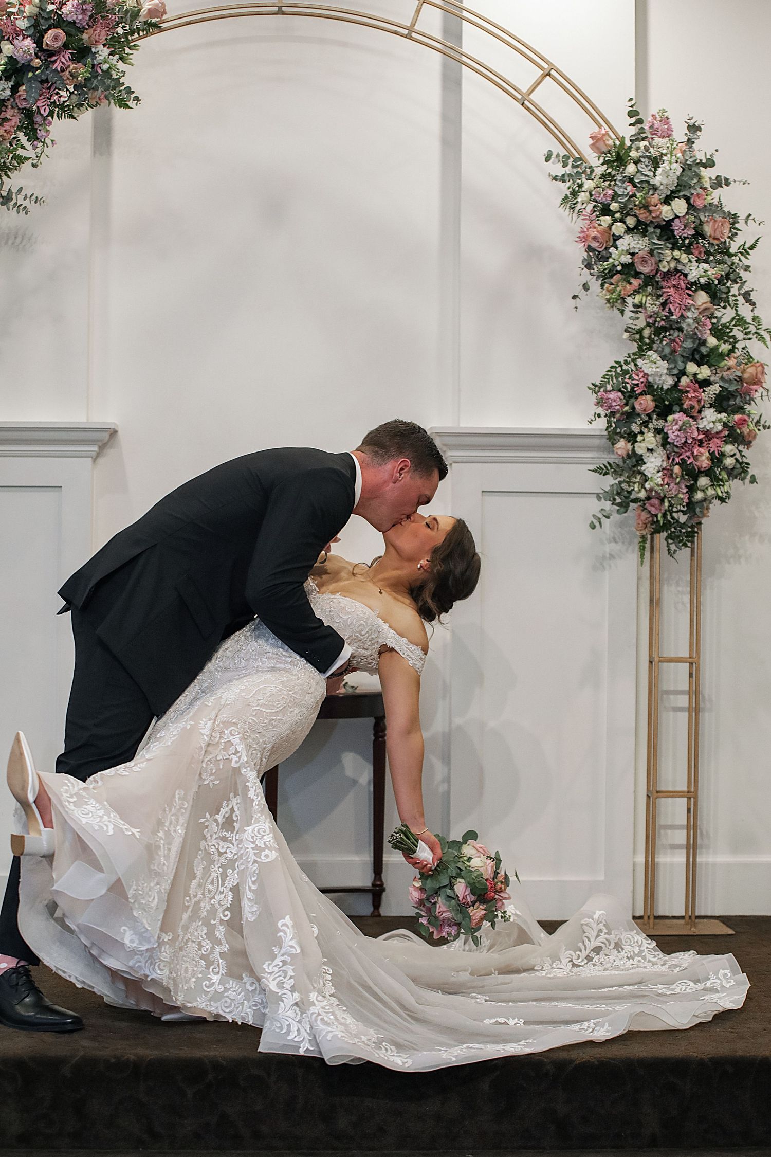 Romantic wedding couple performs dramatic dip kiss pose under floral arch in elegant venue.