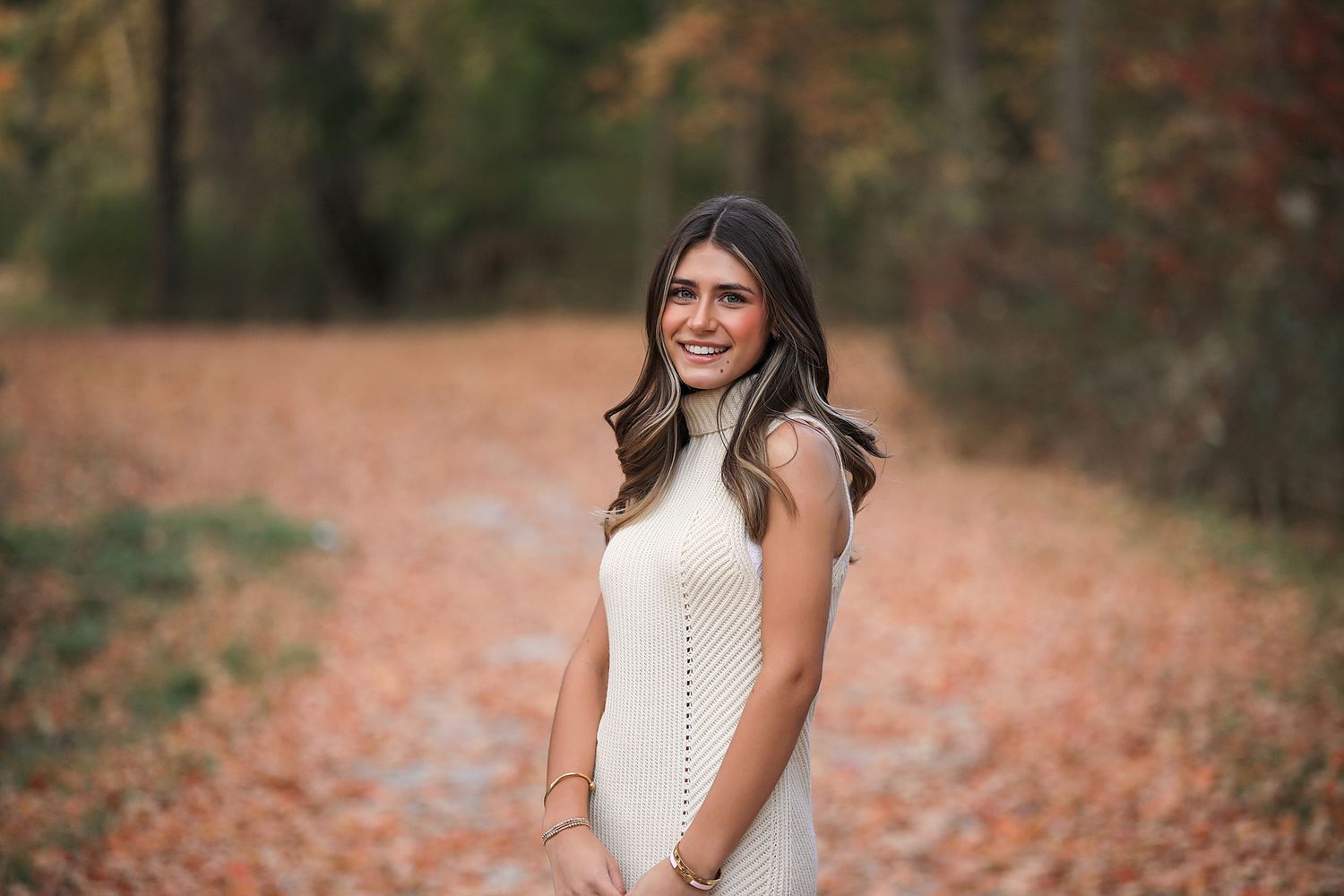 A woman in a white sleeveless dress stands on an autumn path covered with orange fallen leaves.
