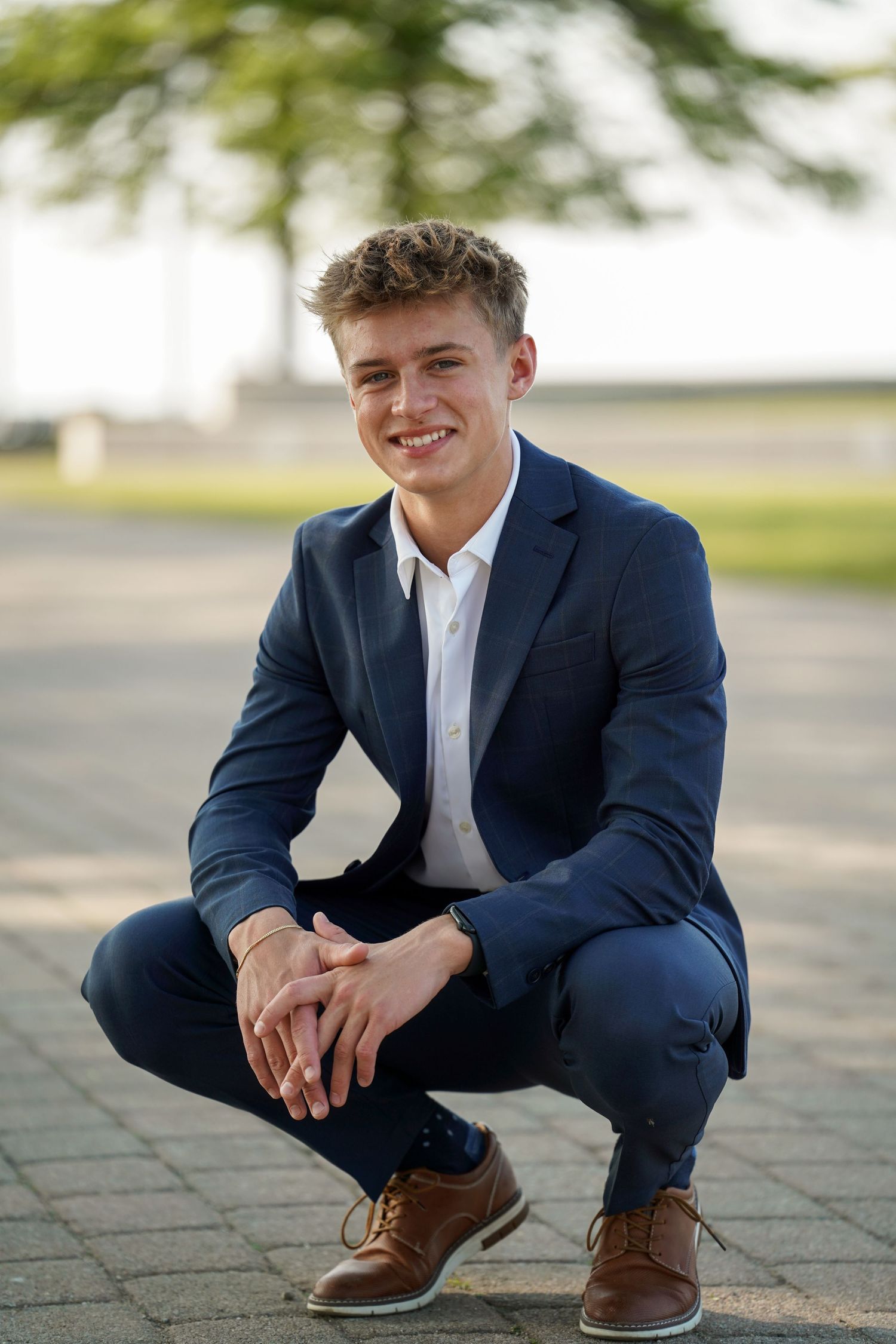 Young person in navy suit and white shirt crouches outdoors in professional portrait pose with brown dress shoes.