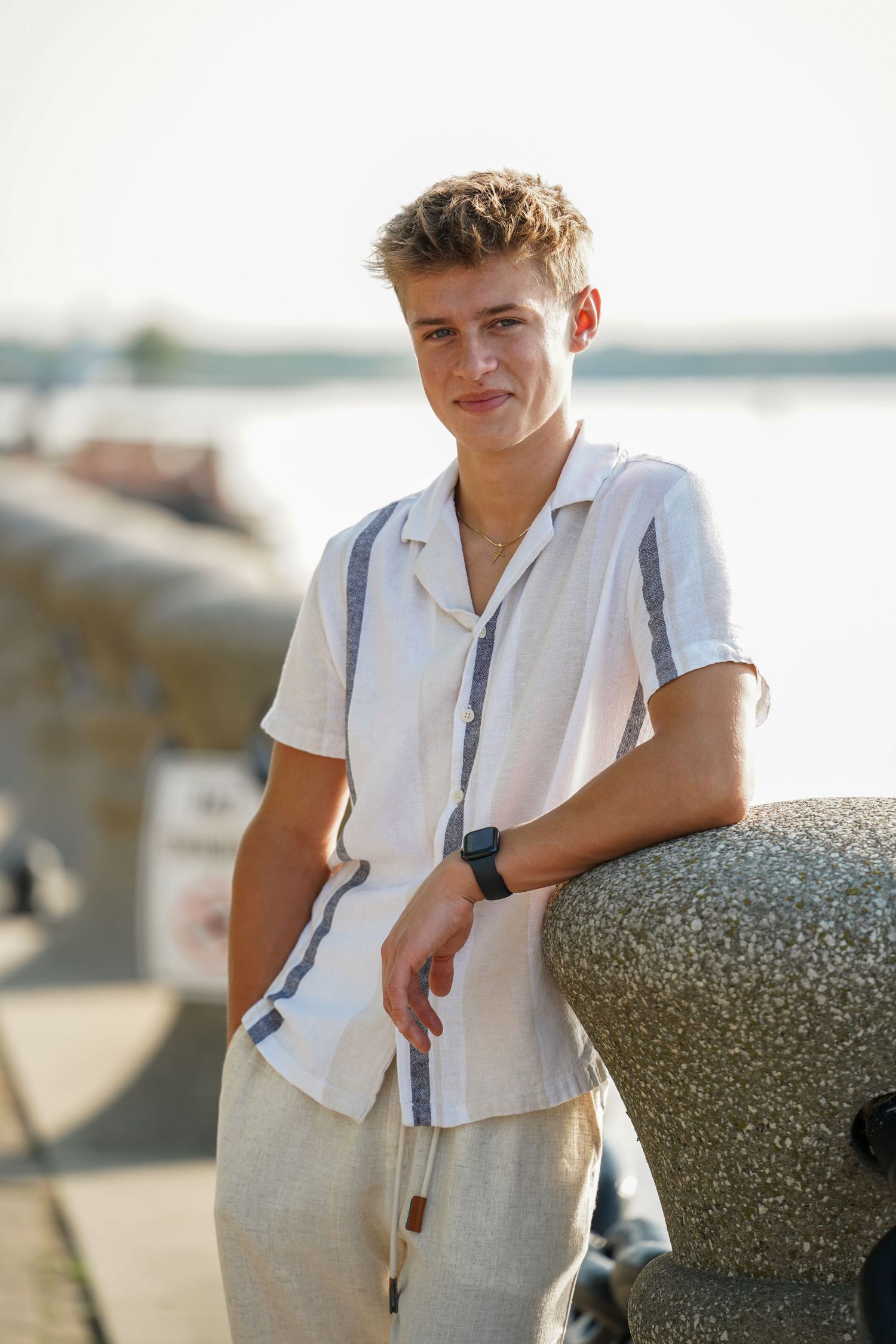 Person in summer attire poses against a lakeside backdrop while wearing a white button-down shirt and light pants.