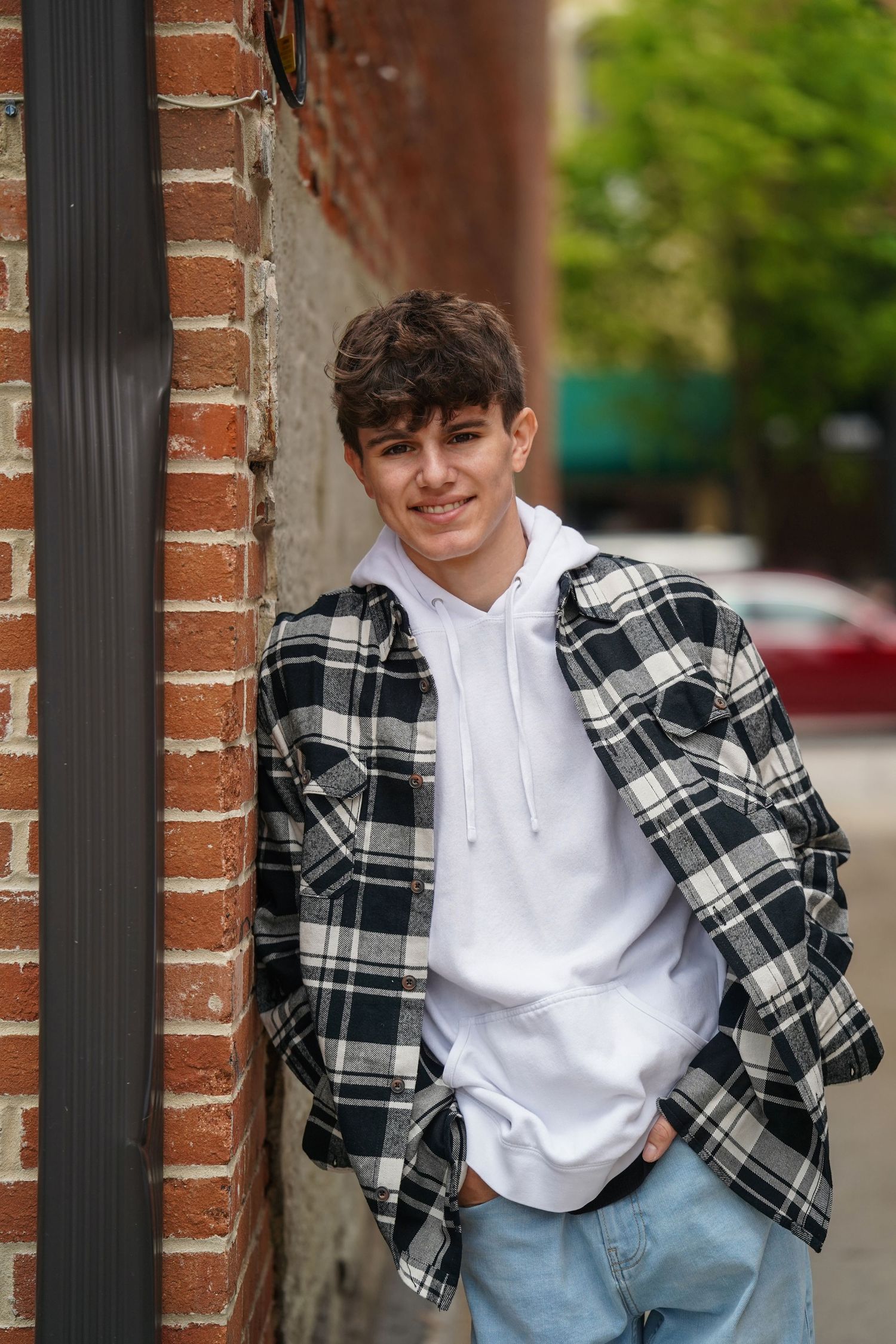 A person in a black and white plaid flannel and white shirt leans against a brick wall while smiling at the camera.