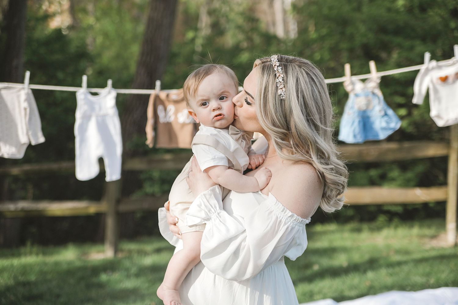 A white dress-clad figure holds a baby in front of a rustic clothesline with hanging laundry in a sunny outdoor setting.