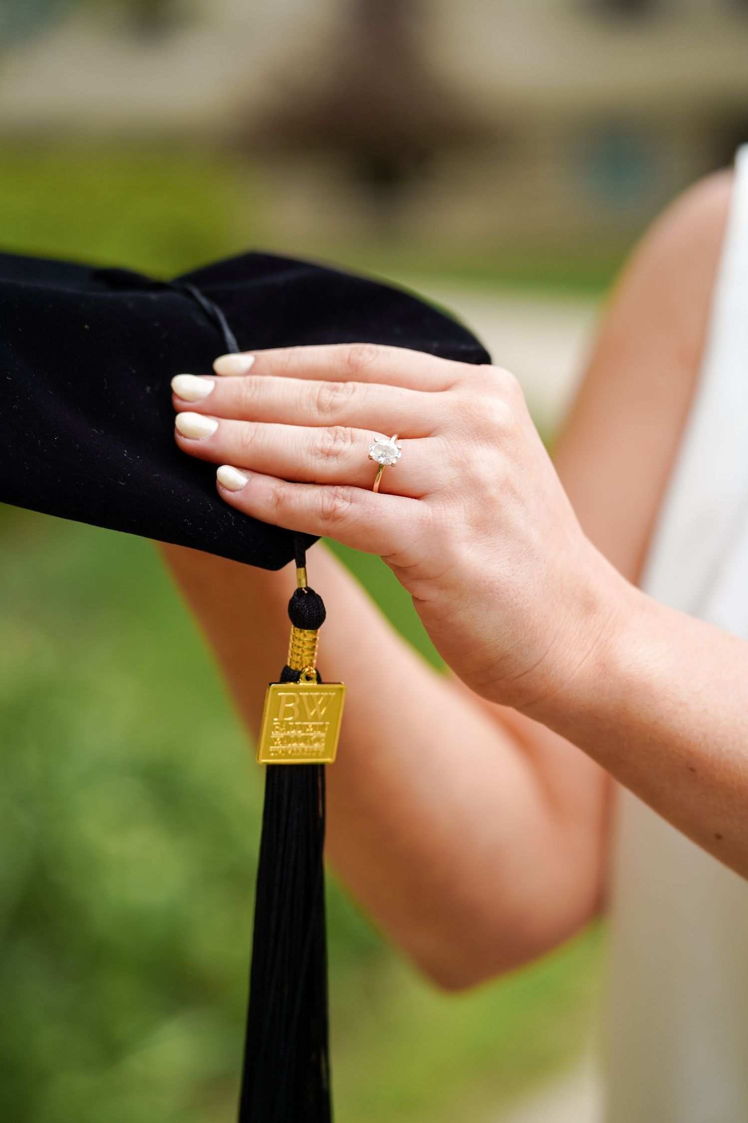 A close-up of hands showing an engagement ring while holding a black graduation tassel with gold detailing.