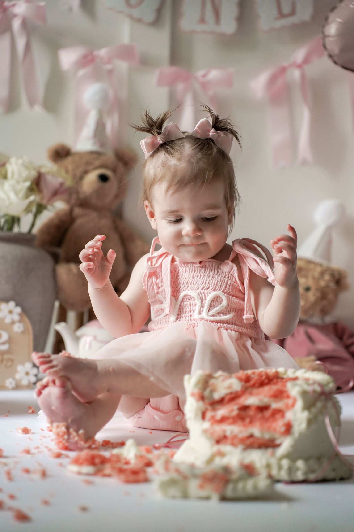 A pink and white smash cake party scene with decorative bows, teddy bears, and scattered cake crumbs on the table.