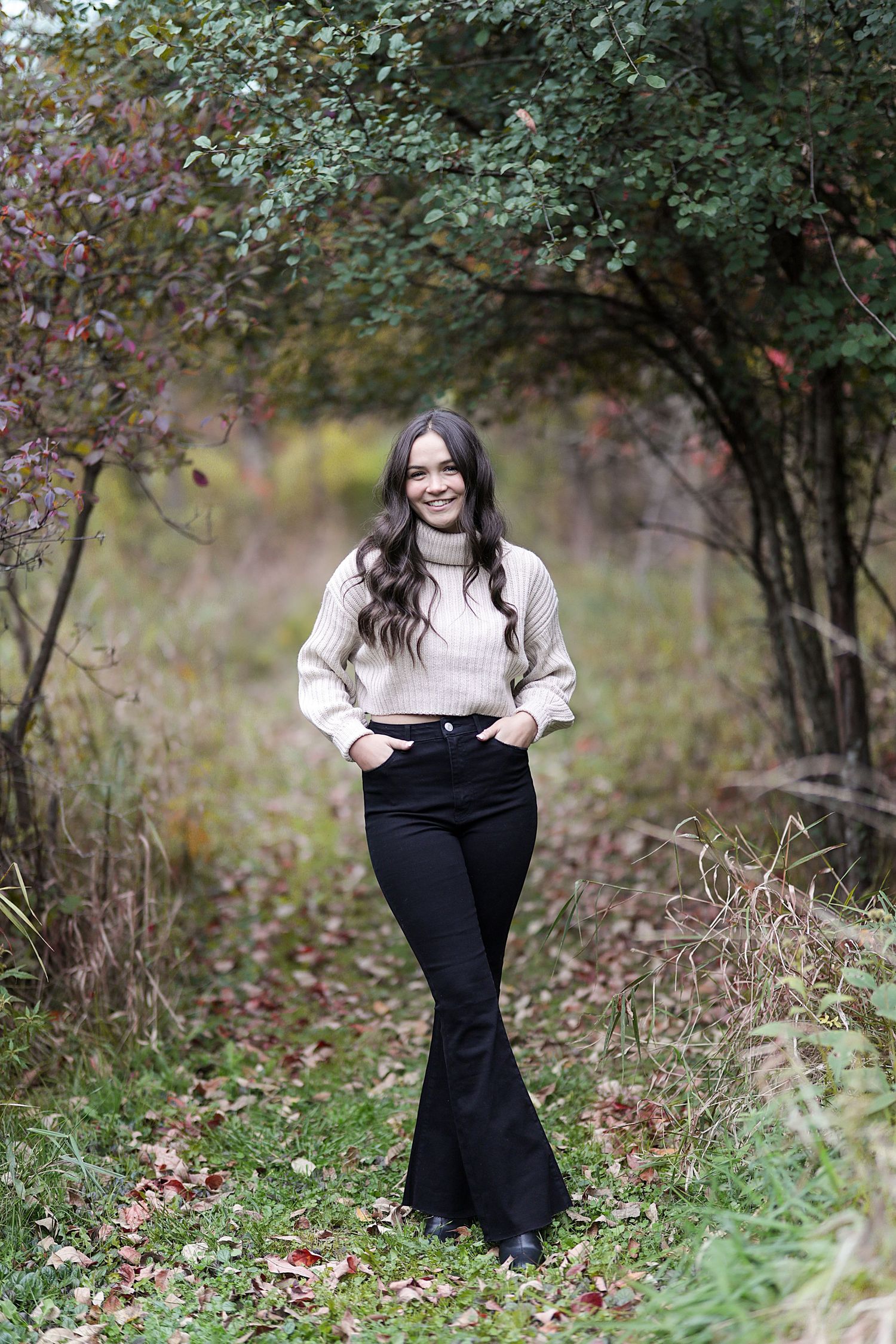 A woman in a cream sweater and black flared pants stands on a nature path surrounded by autumn foliage.