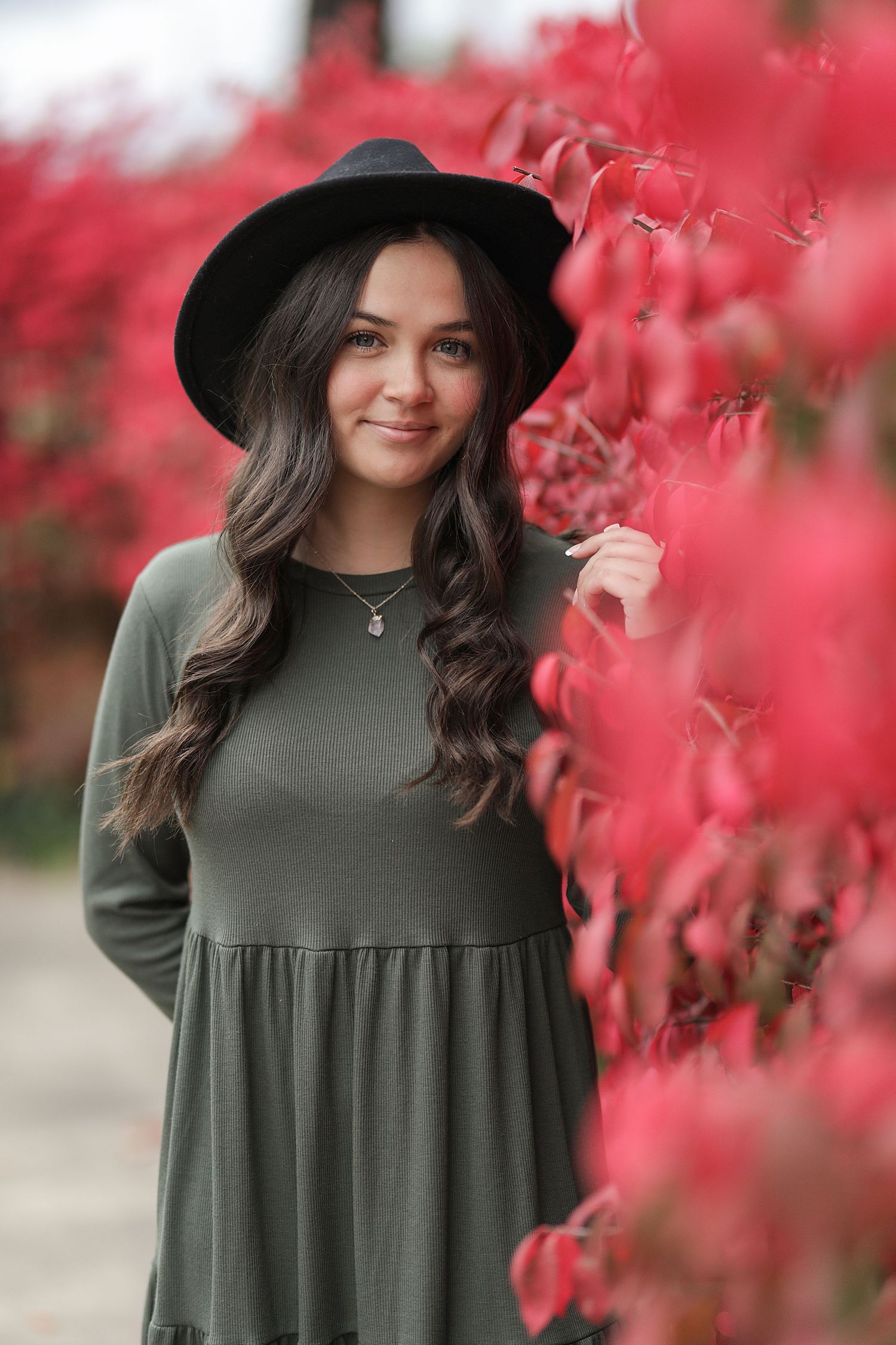 A person in a black hat and olive green dress poses in front of vibrant red autumn foliage.