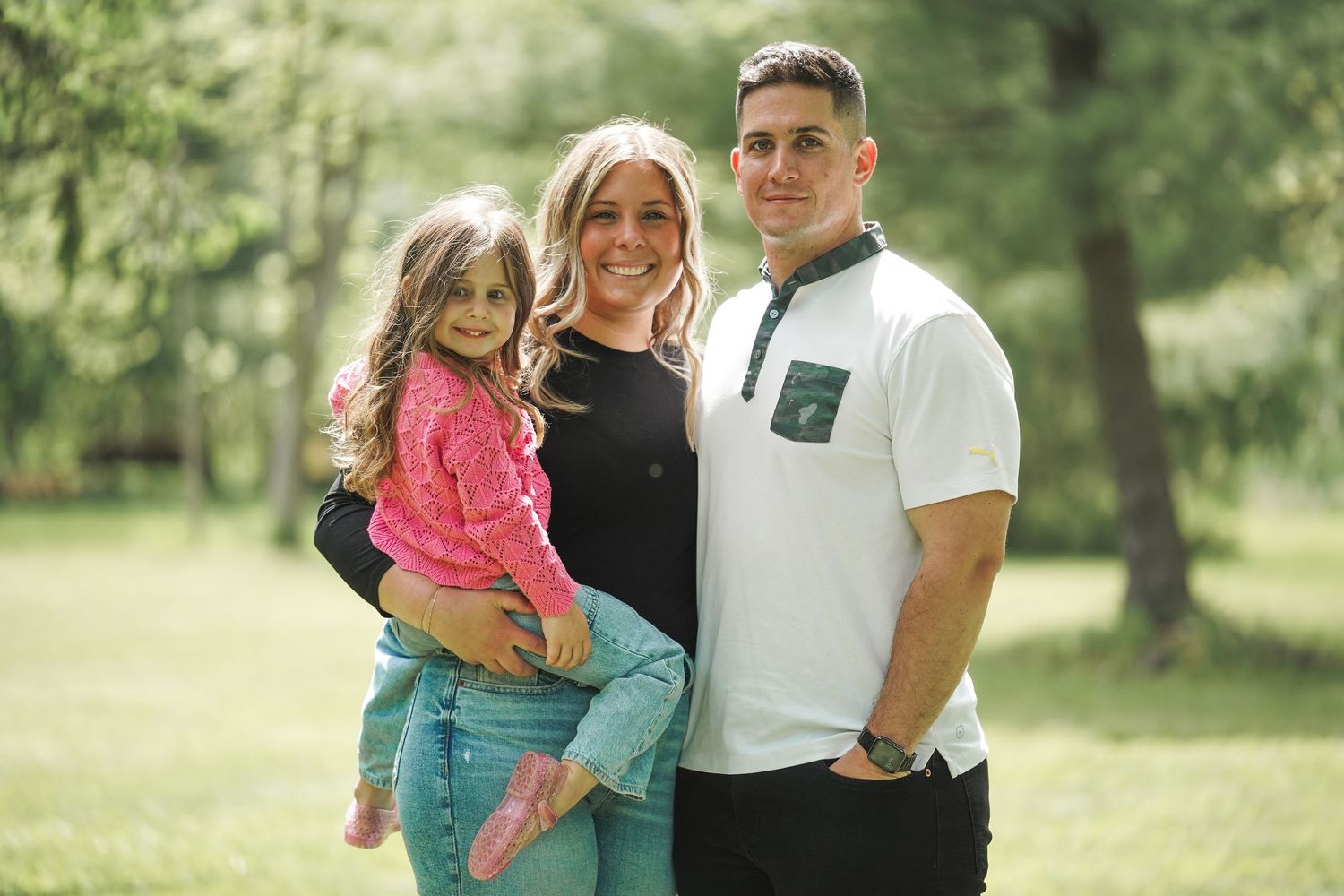 A group of three people standing together outdoors in a sunny park with green trees in the background.