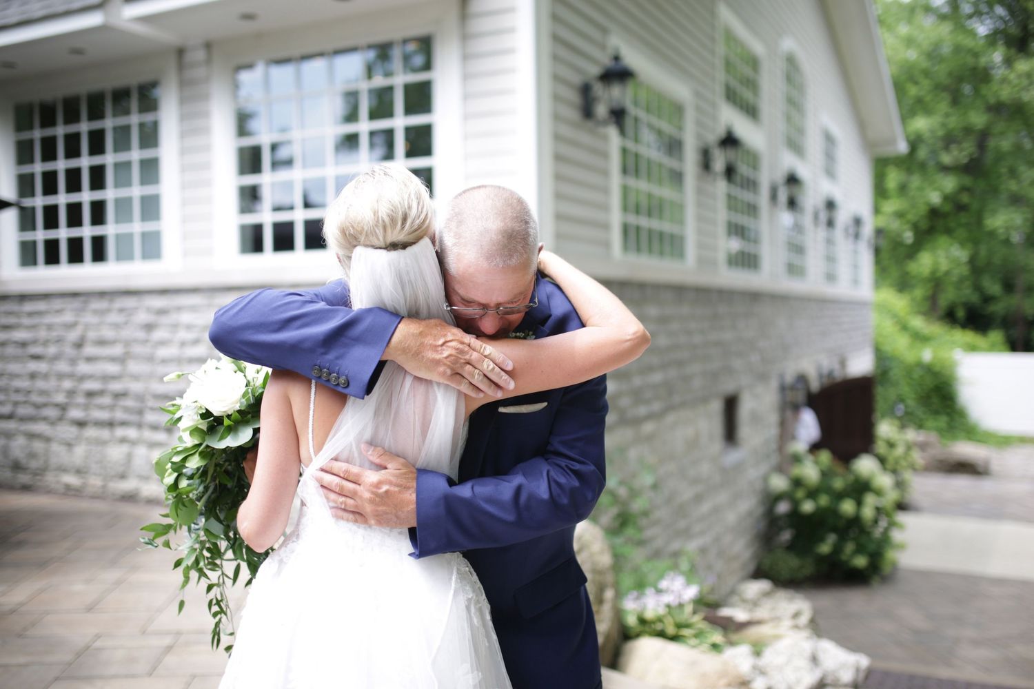 An intimate wedding moment captured outside a white stone building with large windows and flowering bushes.