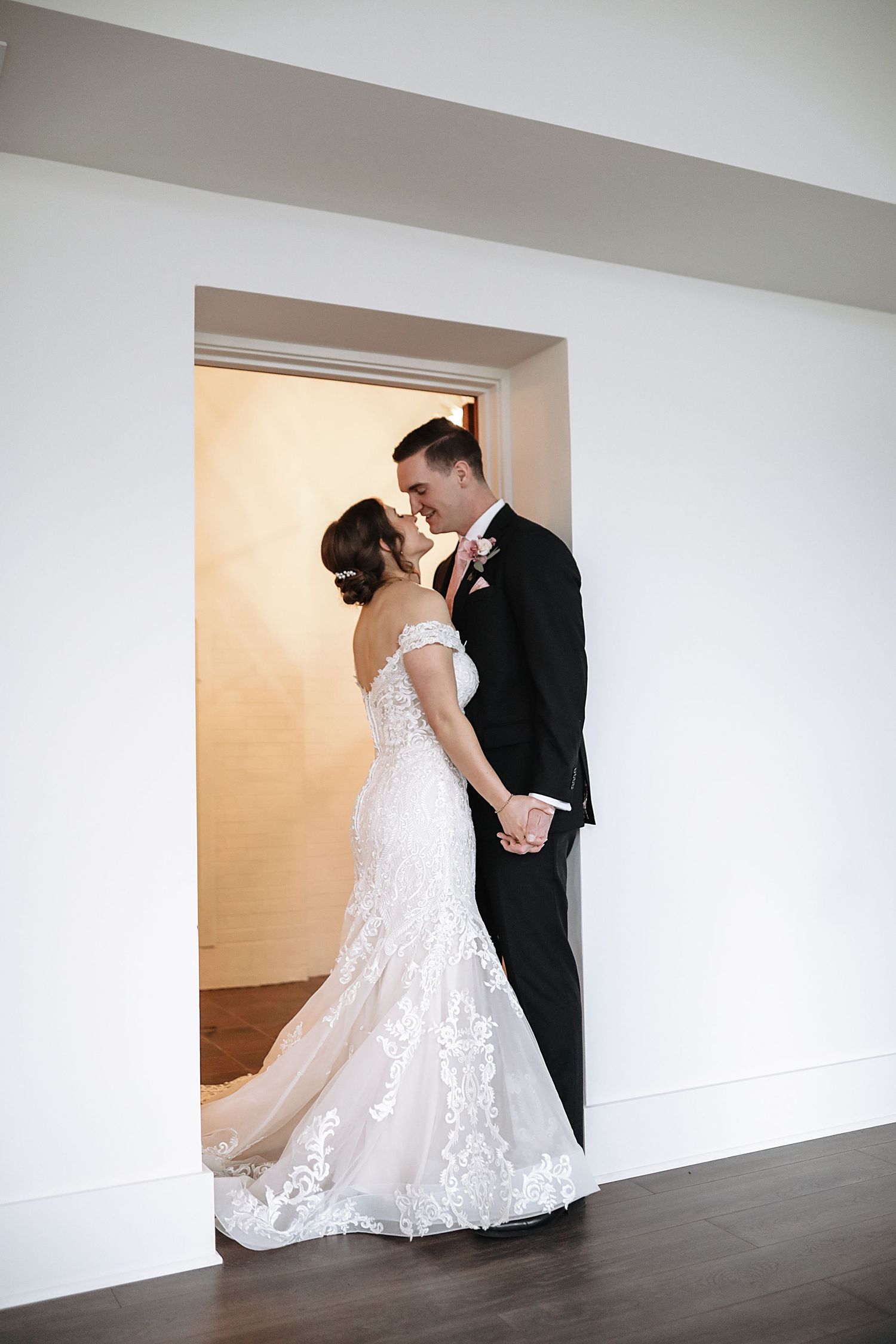 A couple shares a romantic kiss in a doorway, with the bride wearing a lace mermaid gown and groom in classic black tuxedo.