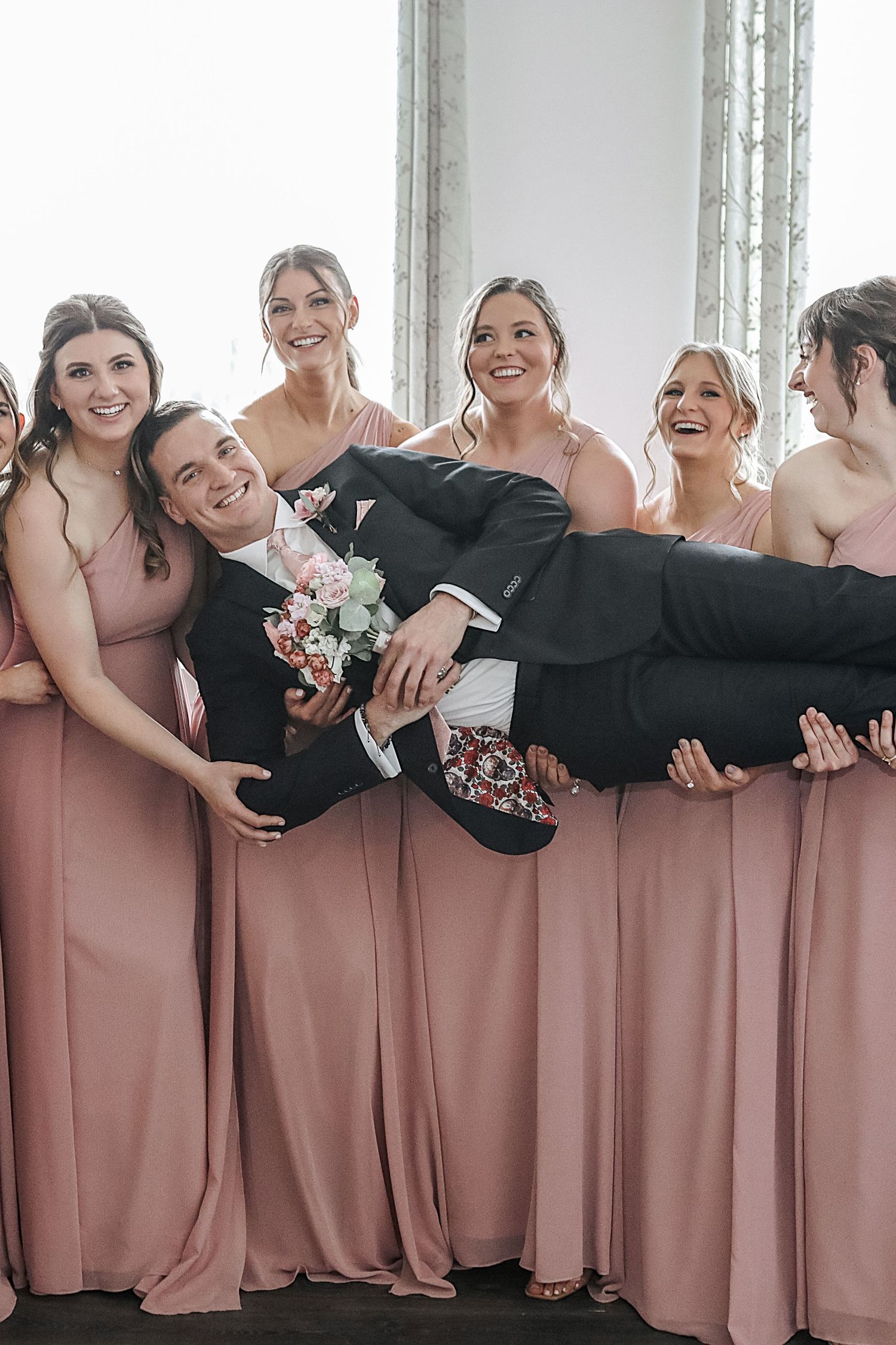 Bridesmaids in dusty pink dresses and black shawls pose together in a playful group photo at a wedding.