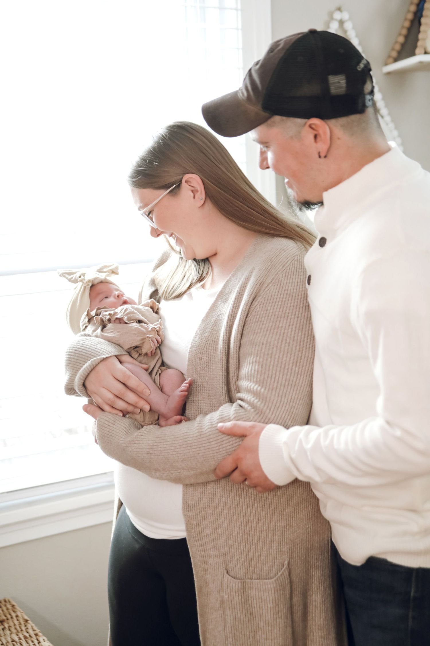A couple in beige and white clothing shares tender moments while holding their newborn baby near a window.